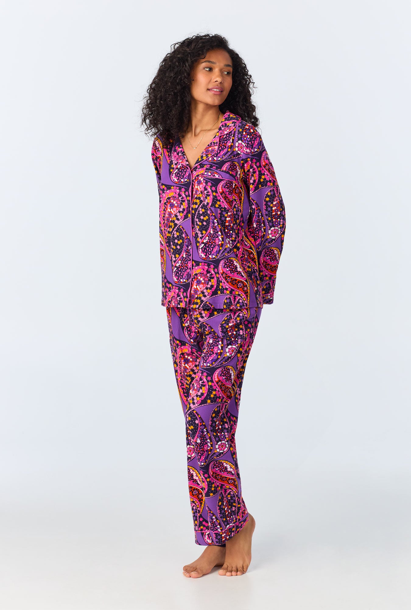 A person with curly hair stands barefoot against a plain light background, wearing the Tangled Paisley Womens Long Sleeve Long Pant Jersey PJ Set in purple with vibrant pink, orange, and black paisley patterns.