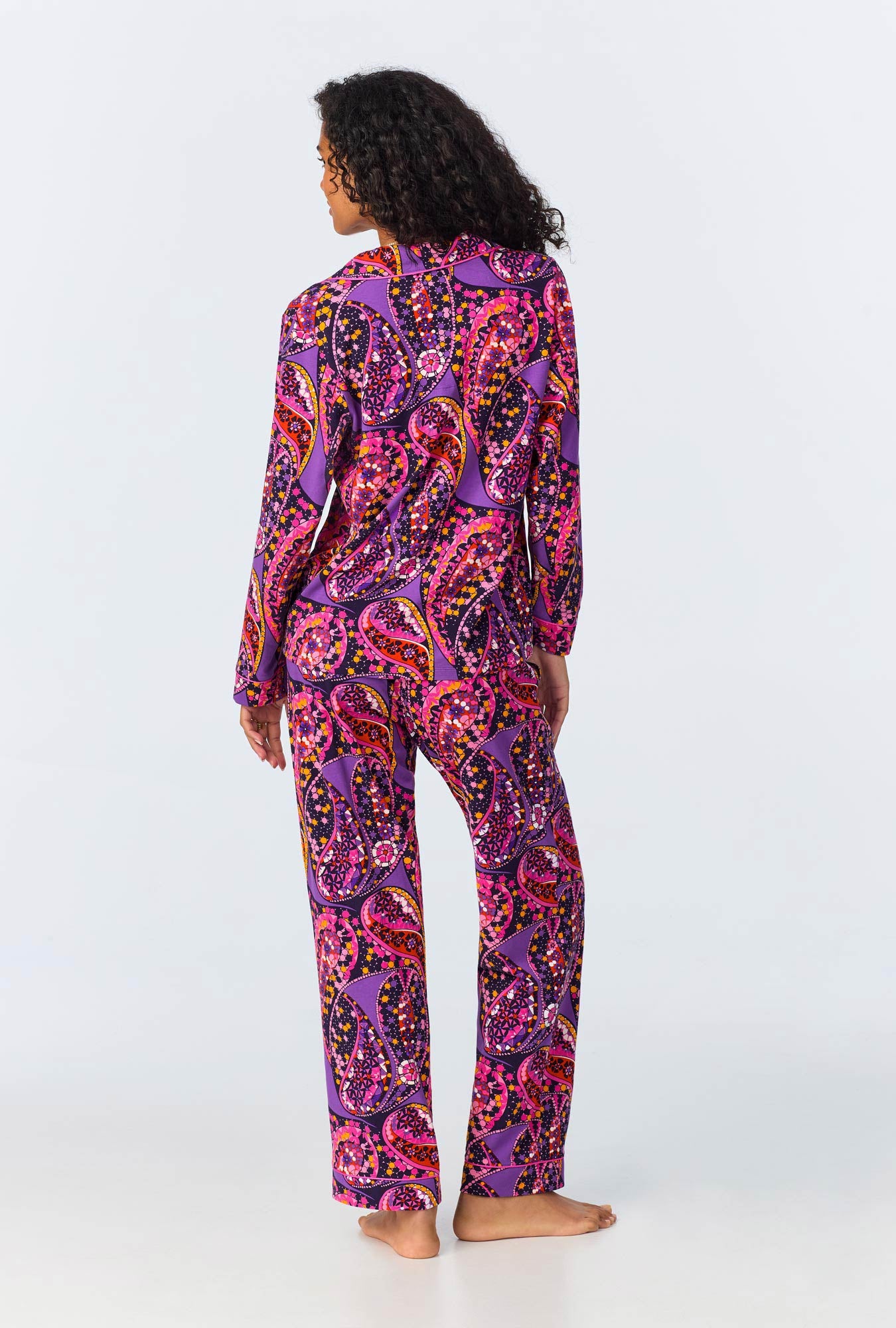 A person with curly hair stands barefoot, facing away, wearing the Tangled Paisley Womens Long Sleeve Long Pant Jersey PJ Set—an eye-catching choice in vibrant purple, pink, and orange paisley on a light background.