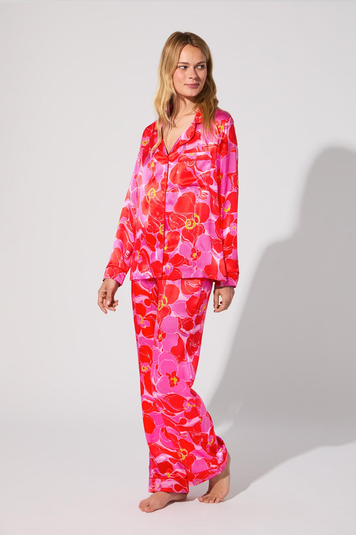 A woman stands barefoot against a plain background, smiling gently while wearing the Silk Classic PJ Set in Winter Orchids—a pink and red floral-patterned satin pajama set with long sleeves and pants.