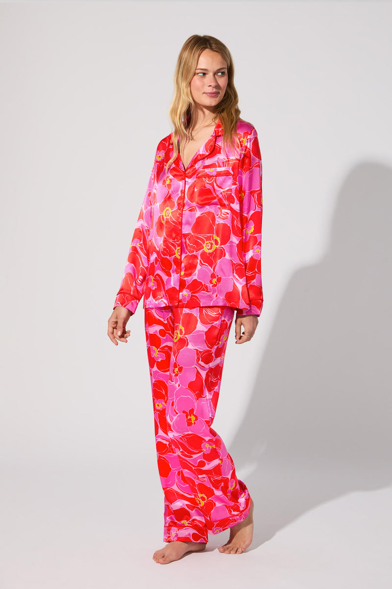 A woman stands barefoot against a plain background, smiling gently while wearing the Silk Classic PJ Set in Winter Orchids—a pink and red floral-patterned satin pajama set with long sleeves and pants.