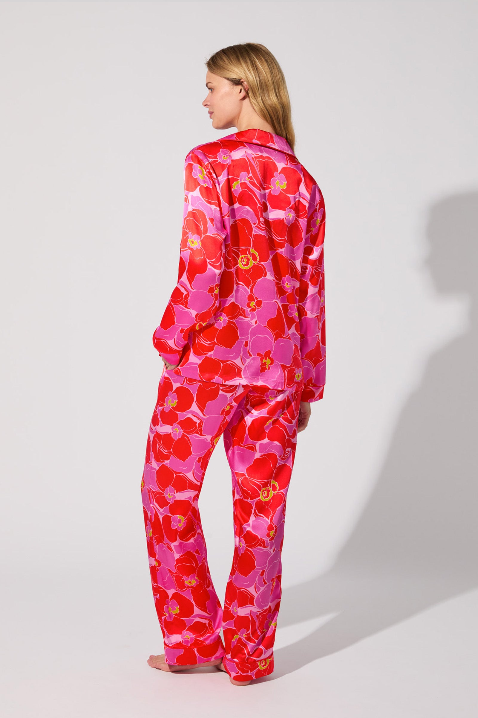 A woman stands barefoot and facing away, wearing the Silk Classic PJ Set - Winter Orchids, a matching long-sleeve pajama set with a vibrant red, pink, and yellow floral pattern on a light background.