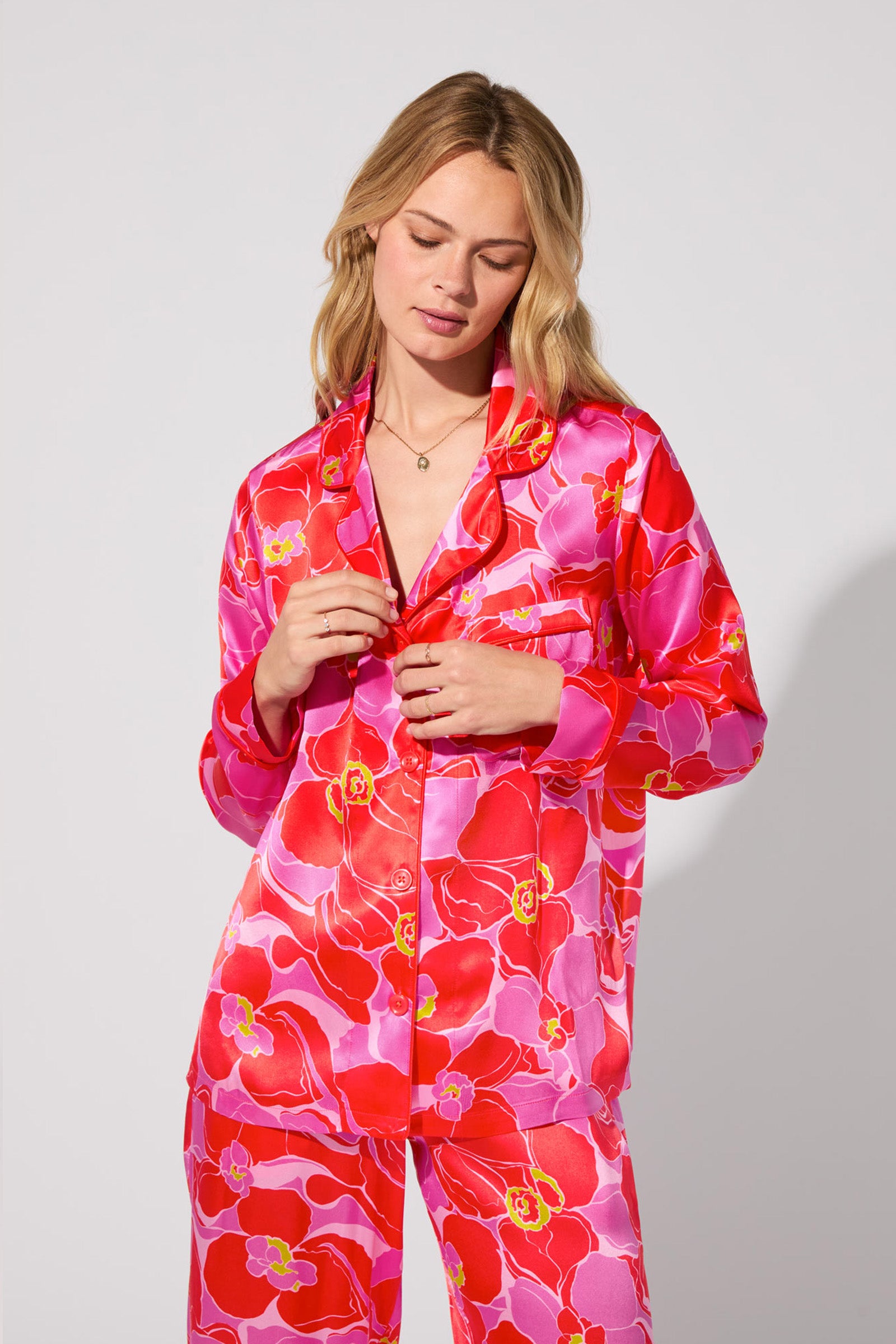 A blonde woman models the Silk Classic PJ Set - Winter Orchids, featuring vibrant pink and red floral patterns, against a light gray background. She looks down slightly, touching the buttons on her shirt.