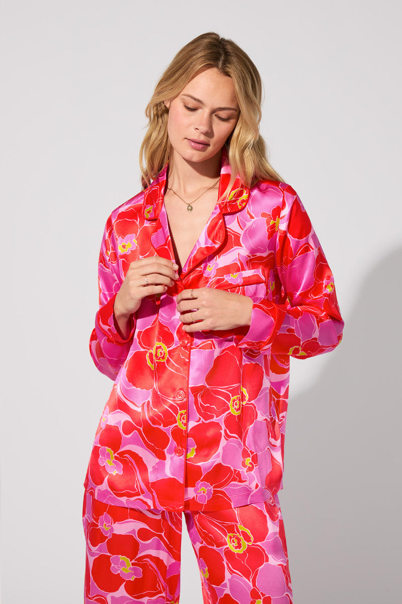 A blonde woman models the Silk Classic PJ Set - Winter Orchids, featuring vibrant pink and red floral patterns, against a light gray background. She looks down slightly, touching the buttons on her shirt.