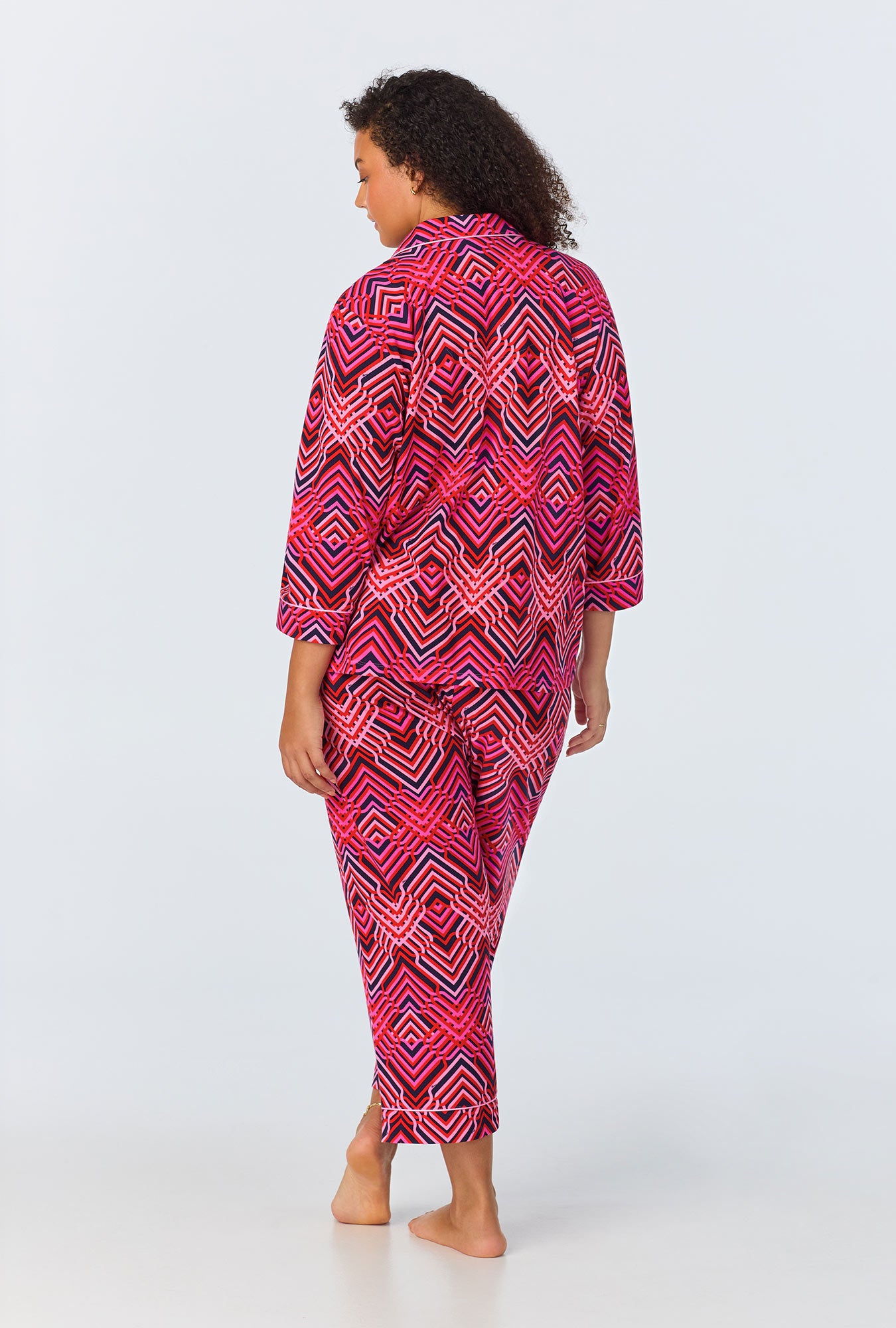 A woman with loose dark curls stands barefoot, facing away in the Geo Trellis Womens 3/4-Sleeve Cropped Pant Jersey PJ Set, featuring a bold red, pink, and white geometric pattern—an ideal pick for womens sleepwear.