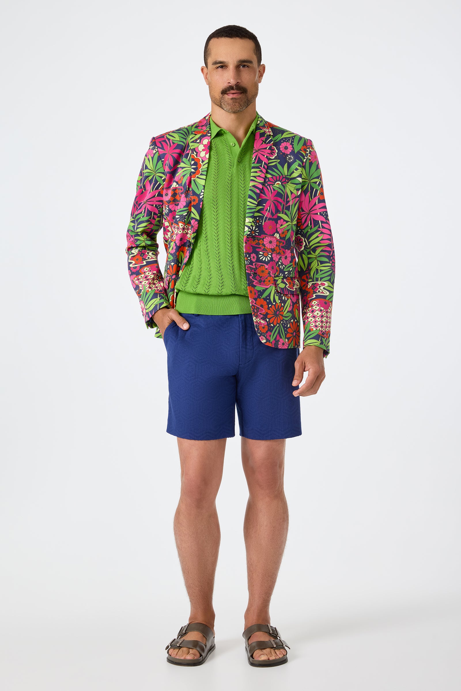 A man with a mustache stands against a white background wearing the Thurston Blazer - Fumi Floral, a bright green sweater, blue shorts, and brown sandals.