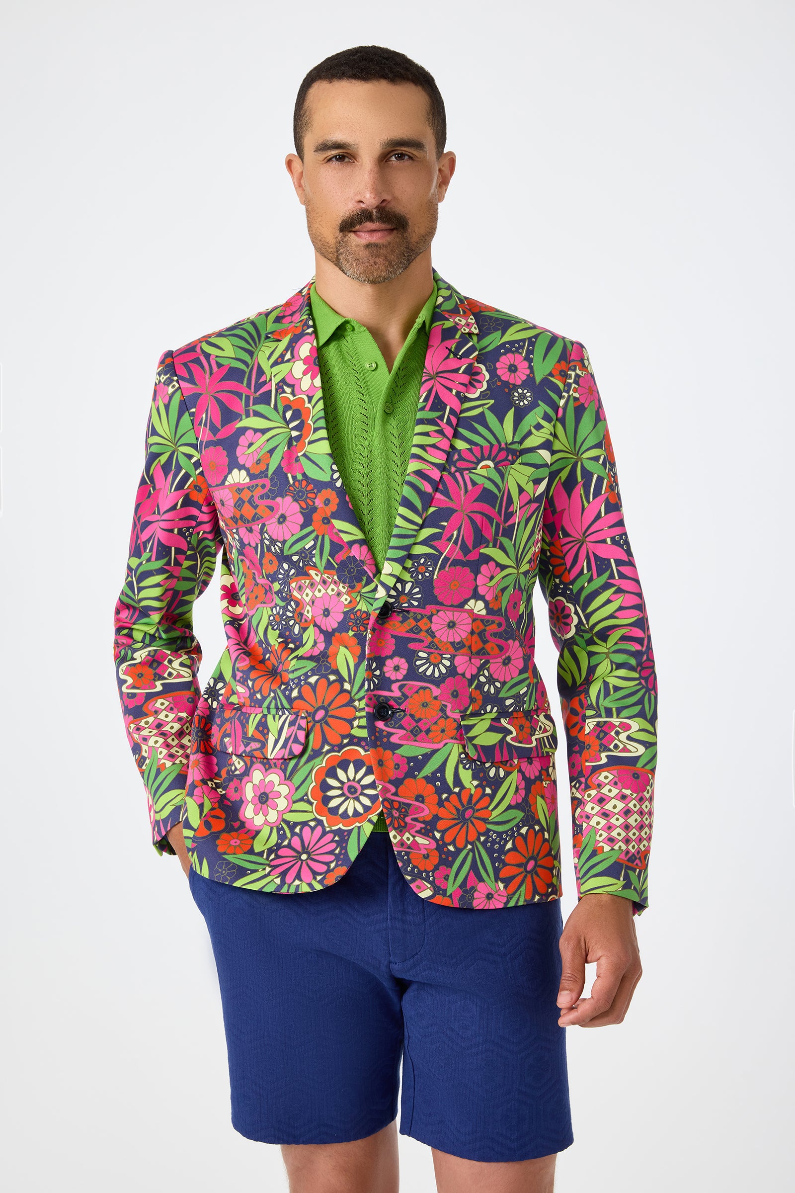 A man stands against a plain white background wearing the Thurston Blazer in Fumi Floral over a green polo shirt and blue shorts.