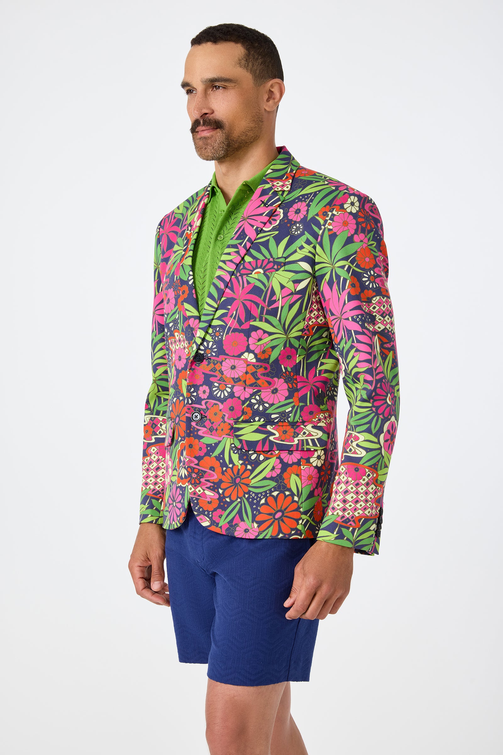 A man with short hair and a mustache stands against a plain background, wearing the Thurston Blazer - Fumi Floral in pink, green, and orange over a green shirt with blue shorts, displaying a neutral expression.