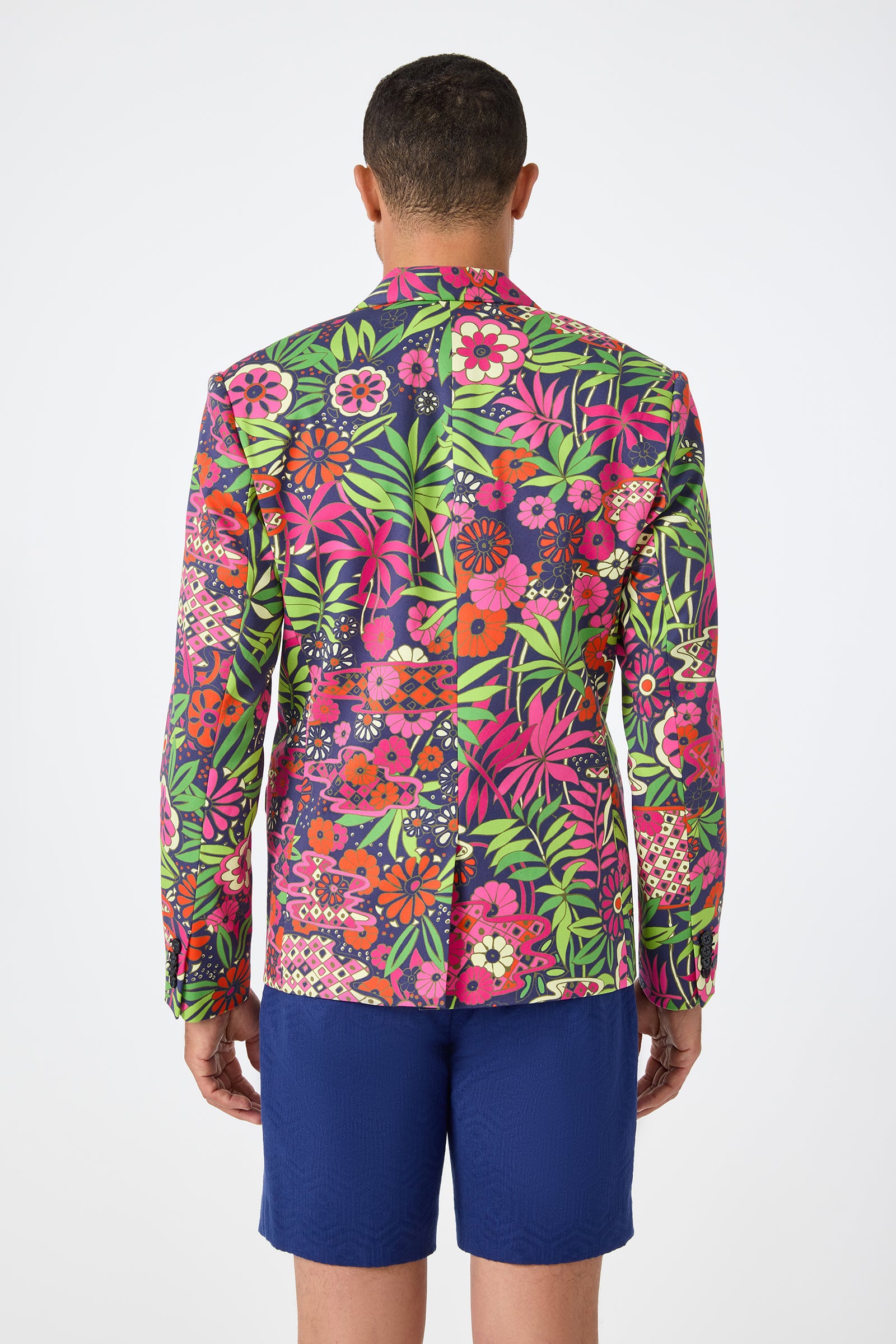 A person faces away, wearing the Thurston Blazer - Fumi Floral, featuring vibrant pink, red, green, and purple floral patterns, paired with solid blue shorts against a plain white background.