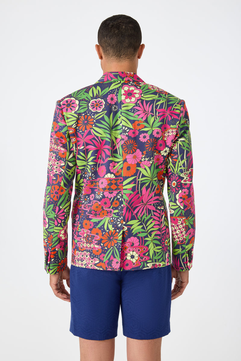 A person faces away, wearing the Thurston Blazer - Fumi Floral, featuring vibrant pink, red, green, and purple floral patterns, paired with solid blue shorts against a plain white background.