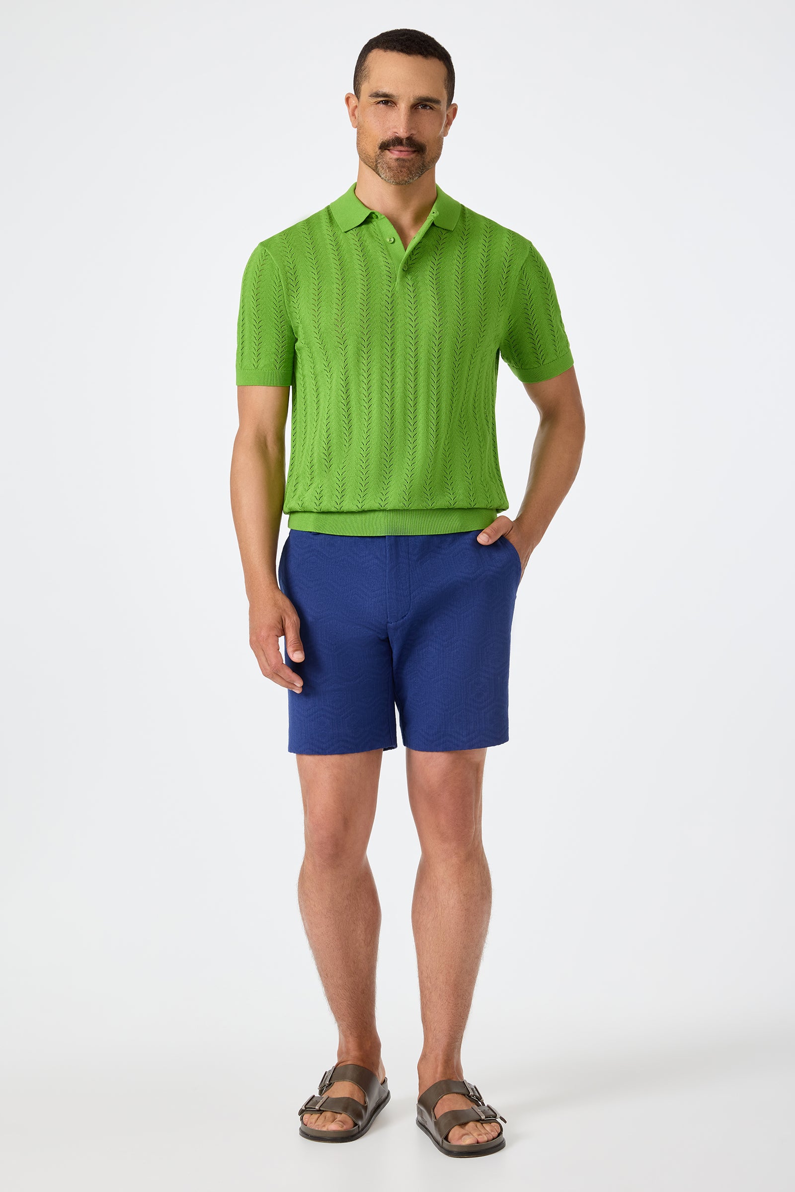 A man with a short mustache and neatly trimmed hair stands against a plain white background, wearing the Cole Short-Sleeve Polo in bright green, blue textured shorts, and brown sandals.