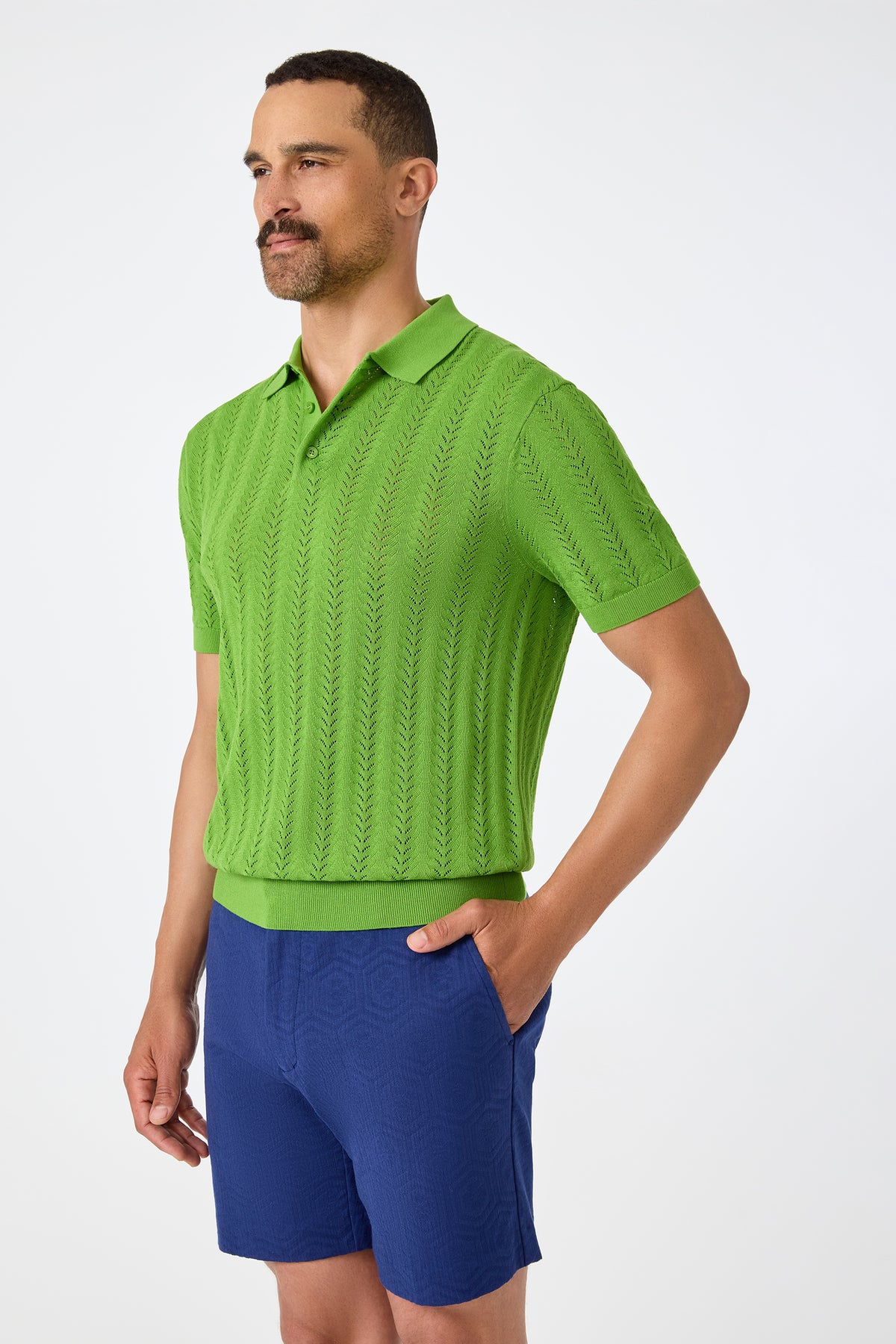 A man with short hair and a beard wears the Cole Short-Sleeve Polo in bright green and blue shorts, standing with one hand in his pocket against a plain white background.