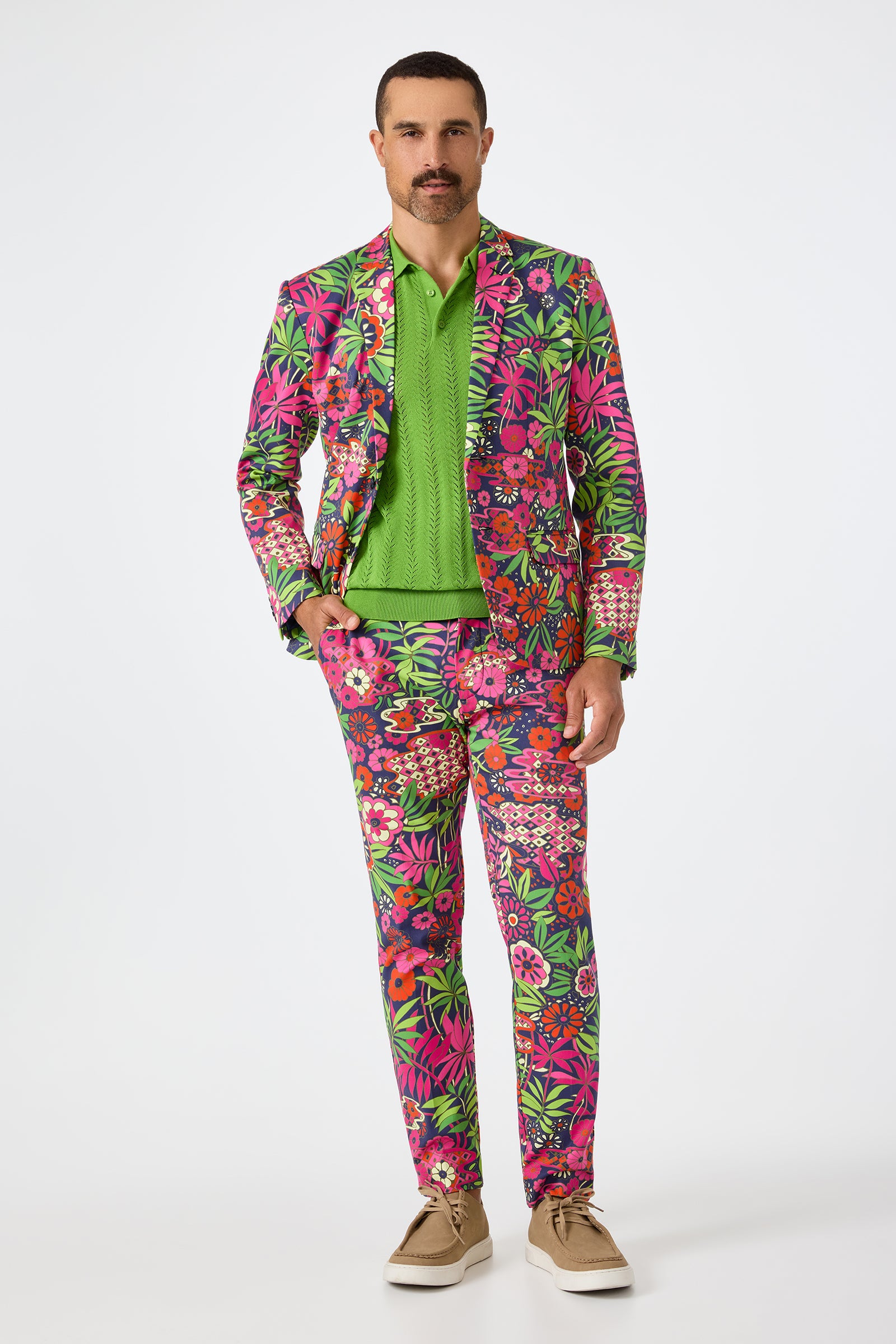A man with short dark hair and a mustache stands facing forward in the Clyde Slim Trouser - Fumi Floral, featuring a vibrant pink, green, and purple floral print, paired with a green shirt and tan shoes. He has a neutral expression.