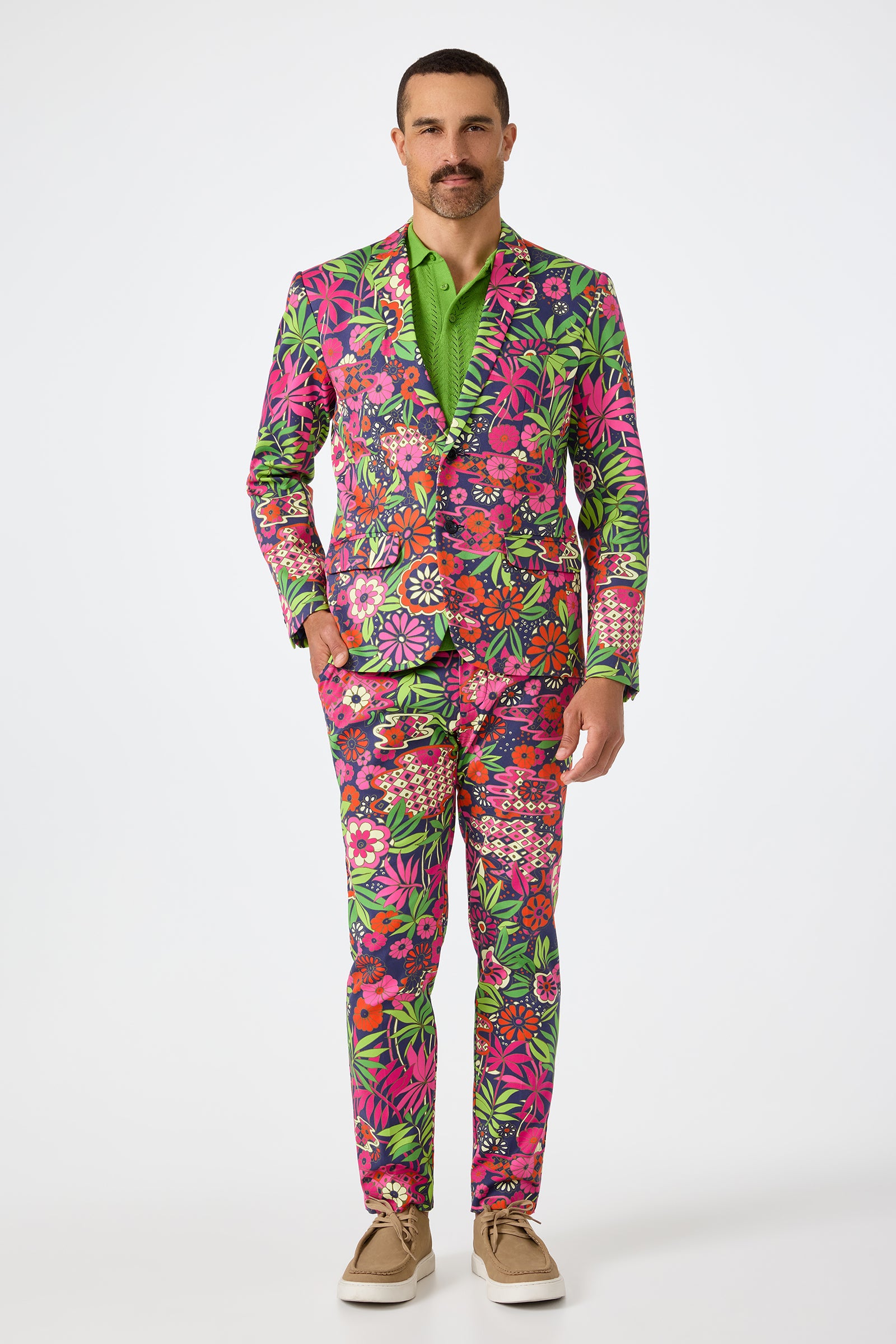 A man stands against a plain background wearing the Thurston Blazer - Fumi Floral, featuring a bold floral pattern in pink, green, red, and white, styled with a bright green shirt and beige shoes.