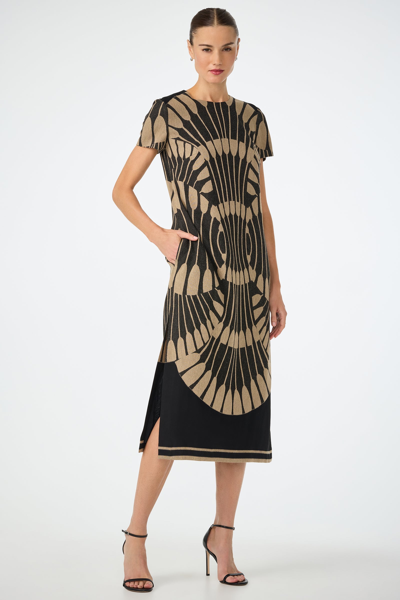 A woman models the Sugarita Midi Dress - Juniper Fil Coupe, featuring bold tan geometric patterns, short sleeves, side slits, and a knee-length fit. She has her hair pulled back and wears black strappy heels while posing with one hand in her pocket.