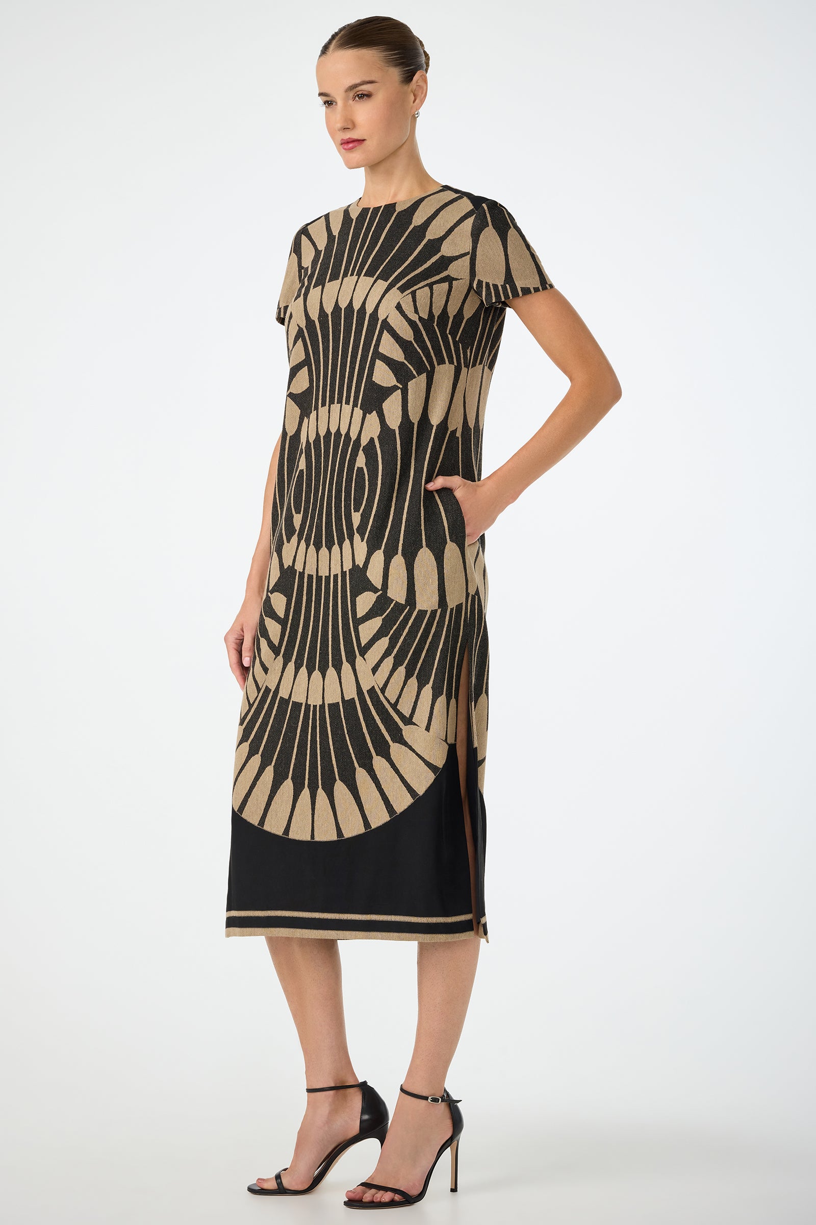 A woman wearing the Sugarita Midi Dress - Juniper Fil Coupe, featuring short sleeves, knee-length fit, bold black and beige geometric pattern with side slit and black hem, paired with black heels against a plain light background.