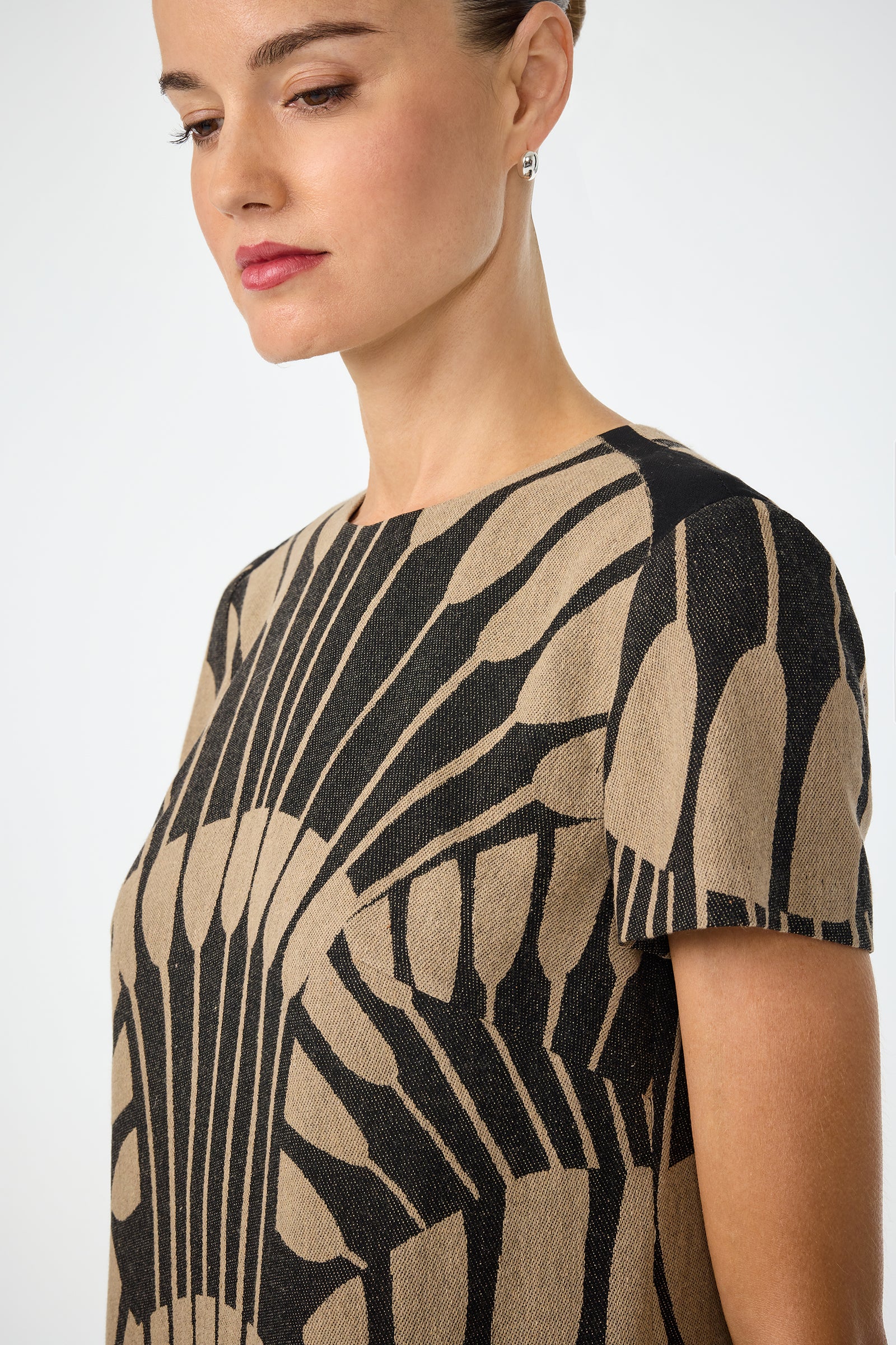 A woman with light skin and brown hair in a bun wears the Sugarita Midi Dress - Juniper Fil Coupe, featuring short sleeves and a bold geometric pattern. She faces slightly left, wearing small hoop earrings with a calm expression.