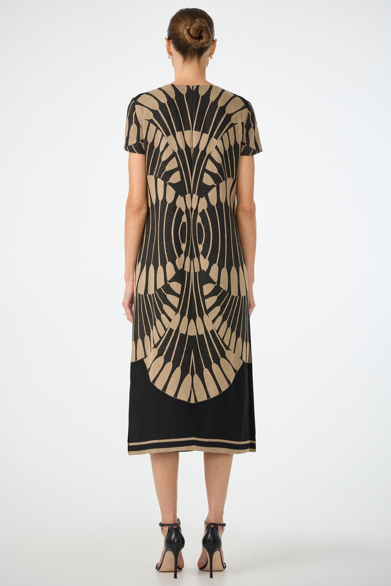 A woman with her hair in a neat bun faces away, wearing the Sugarita Midi Dress - Juniper Fil Coupe, a calf-length black dress with short sleeves, a tan geometric back pattern, and paired with black high-heeled sandals.