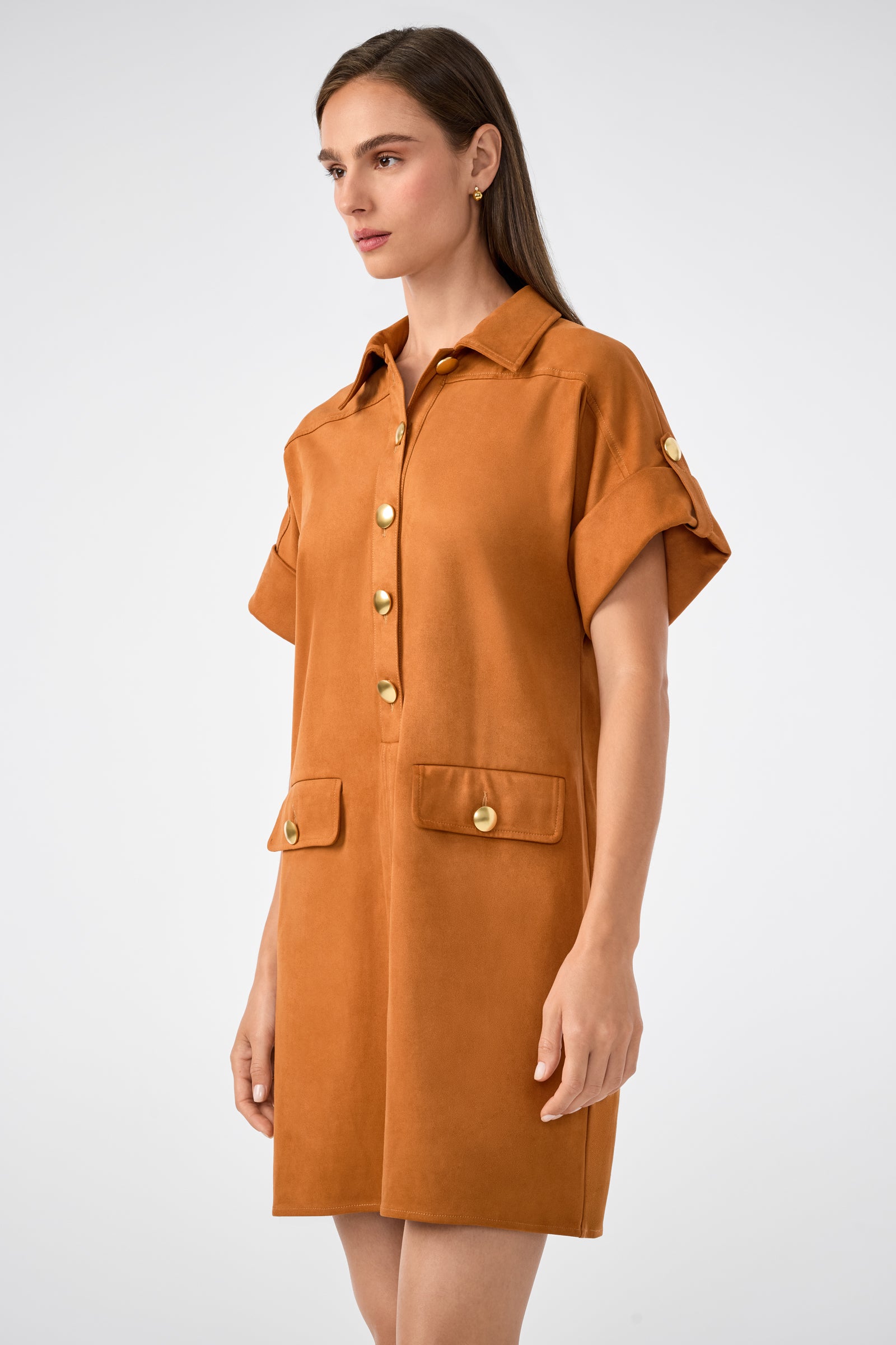 A woman stands against a plain light background wearing the Sequoia Dress, a tan short-sleeved shirt dress featuring gold buttons, front flap pockets, and rolled sleeves.