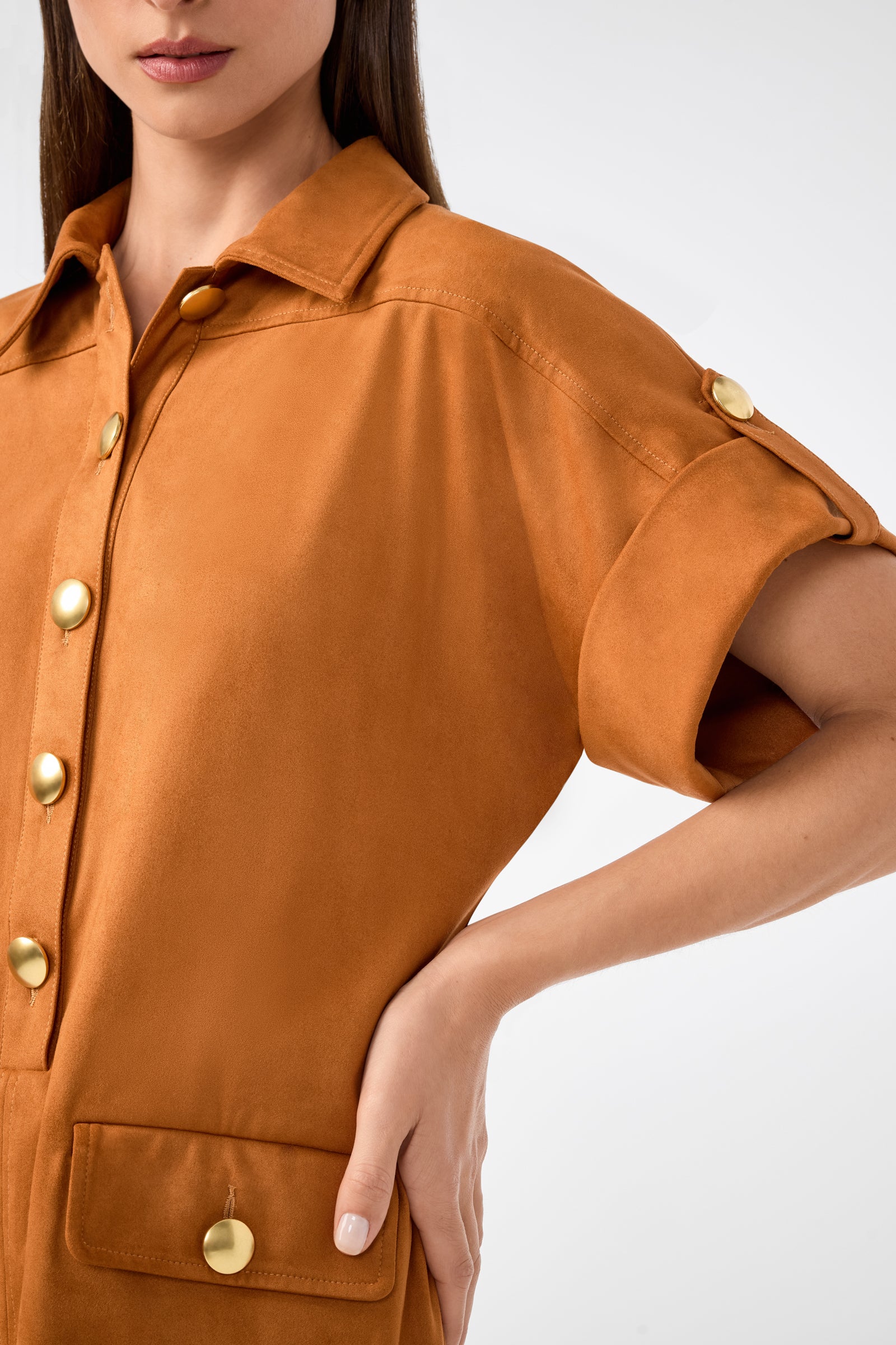 A woman stands against a plain background wearing the Sequoia Dress—a short-sleeved, button-up, camel-colored style with gold buttons and one hand on her hip.