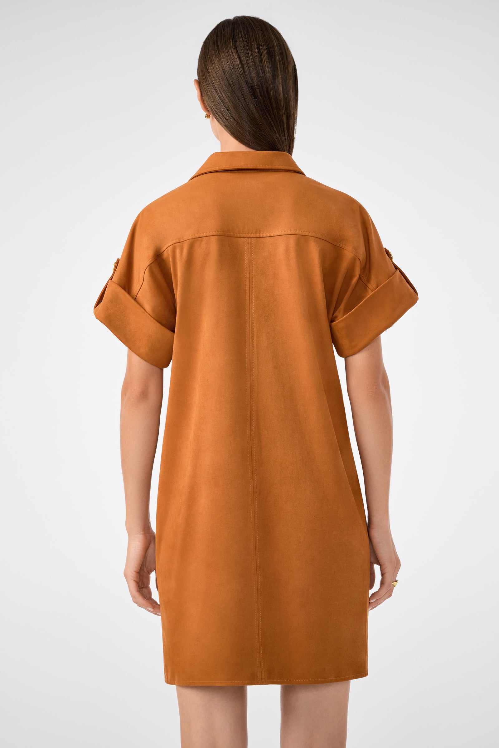 A person with long brown hair is seen from the back wearing the Sequoia Dress—a loose, short-sleeved caramel brown dress with rolled cuffs—against a plain white background.