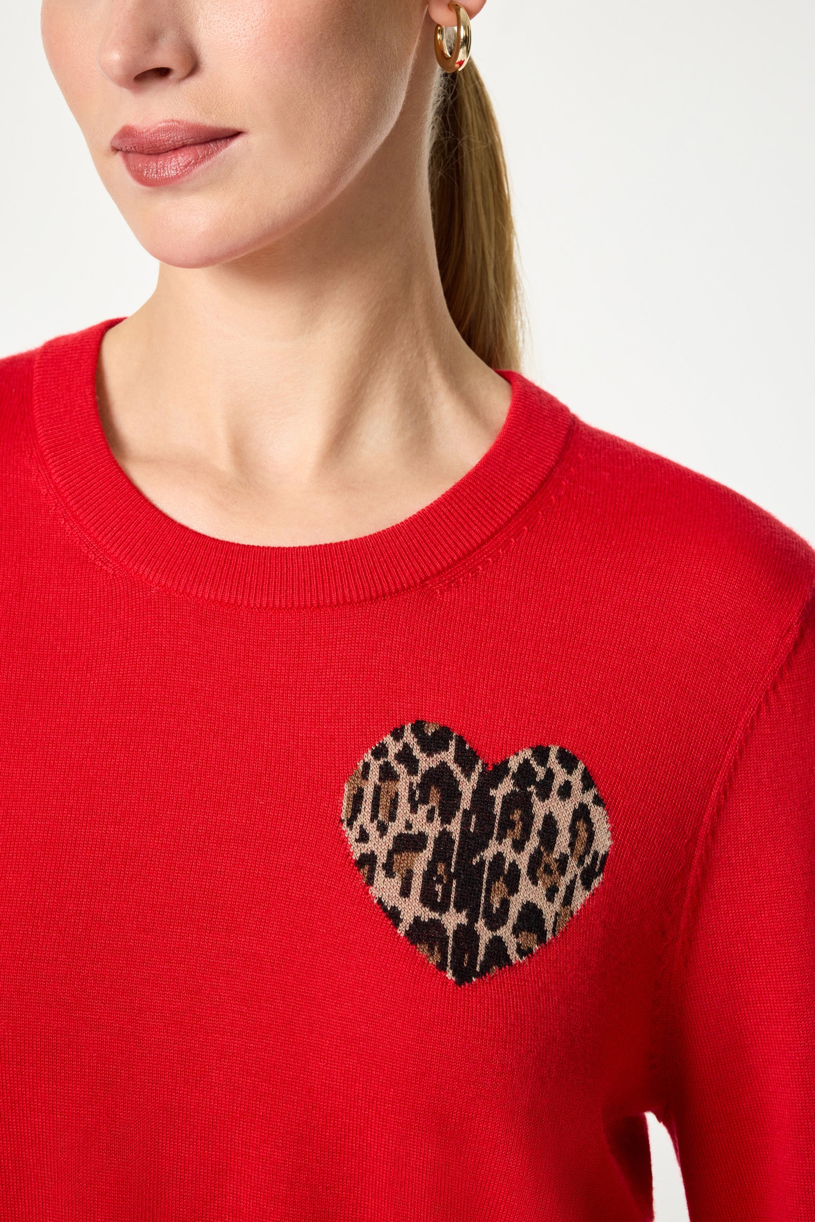 A woman with light skin wears the Jaguar Heart Crewneck Sweater—a bright red top with a leopard-print heart patch on the chest. Only her lower face, neck, and upper torso are visible, and she sports a small gold hoop earring.