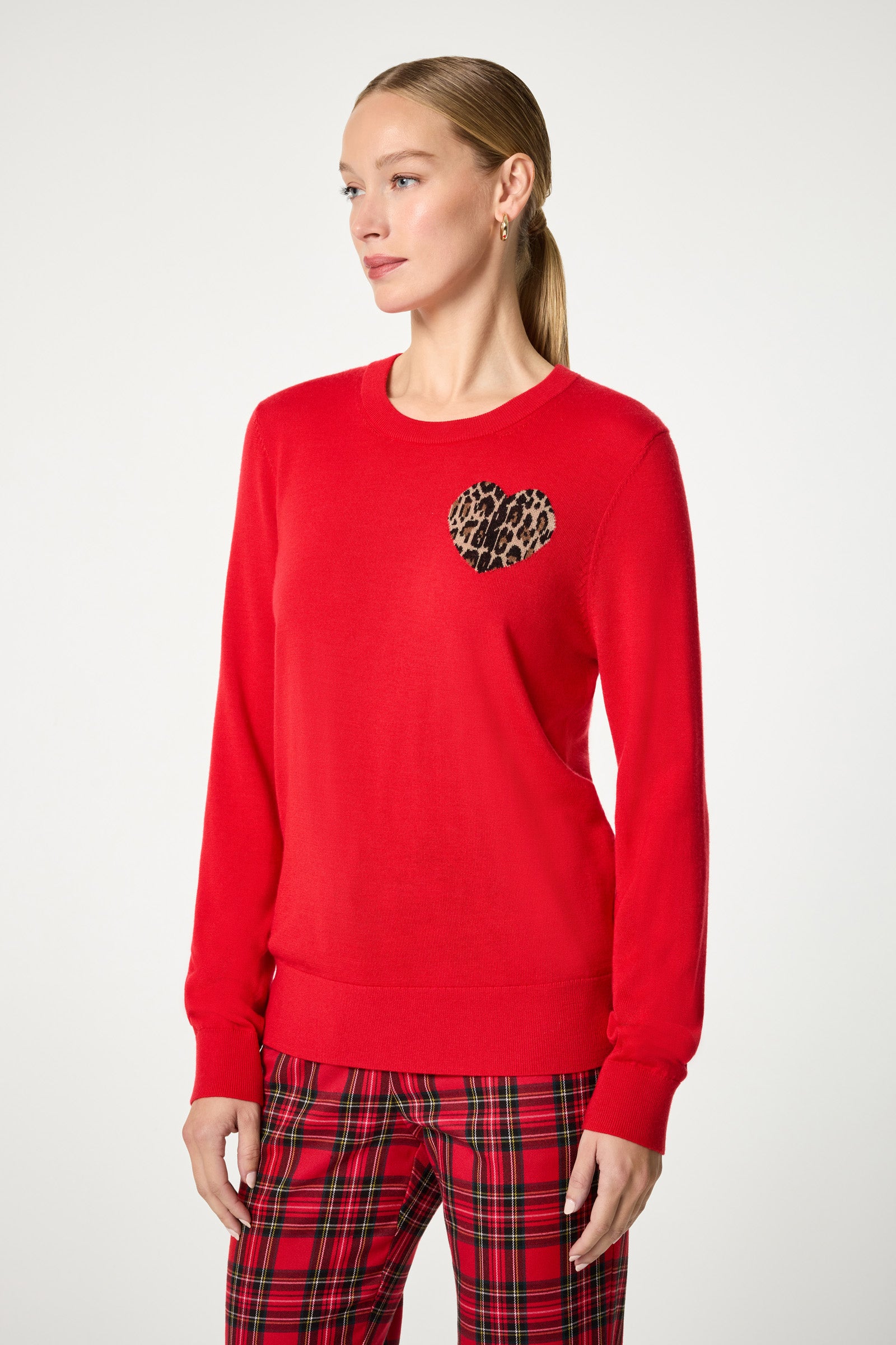 A woman wears the Jaguar Heart Crewneck Sweater, featuring a bold red color and a leopard print heart patch on the chest, paired with red plaid pants, standing against a plain white background.