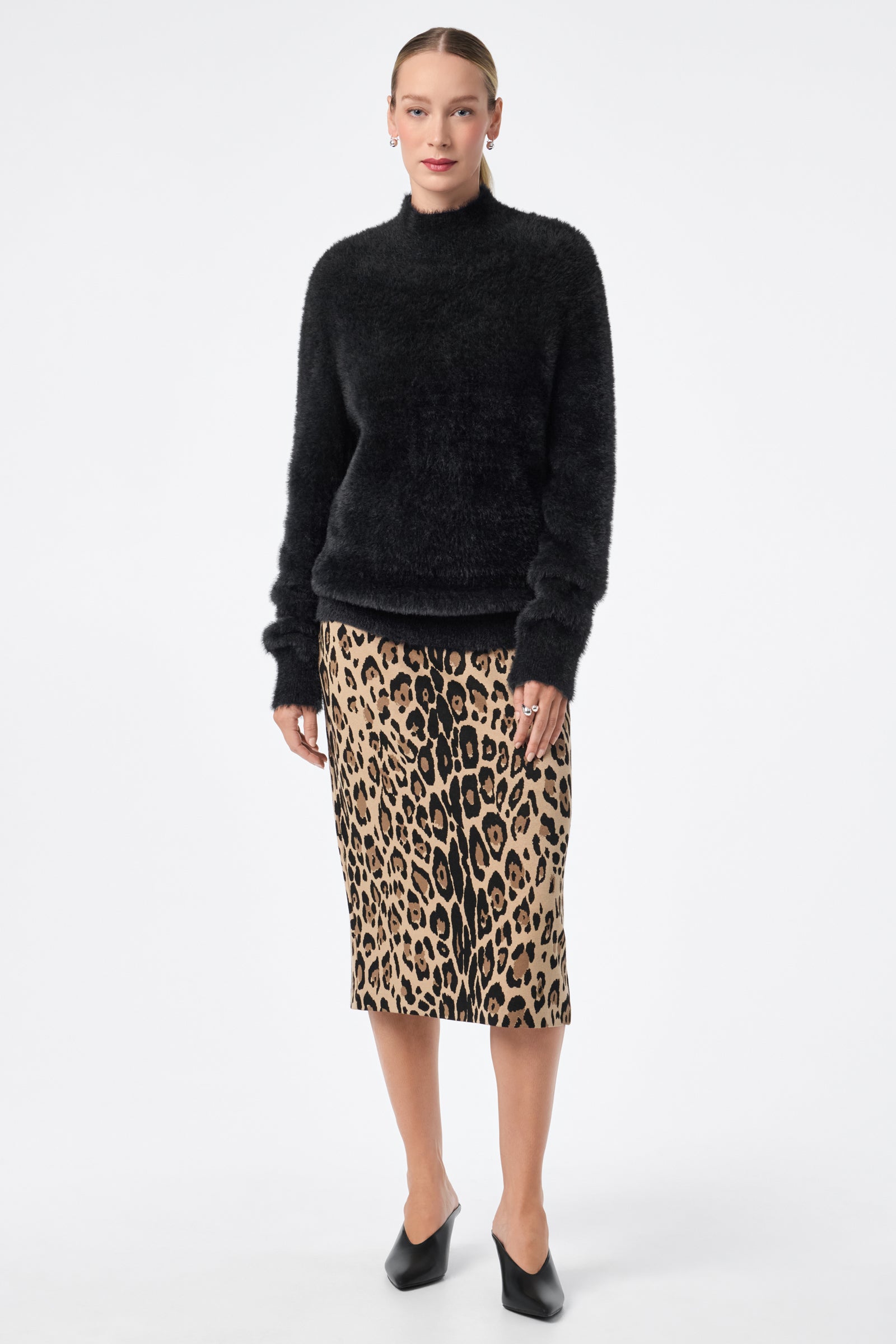 A person stands facing forward in the Dame Dress—a black fuzzy sweater with a knee-length leopard print skirt—paired with black pointed-toe shoes. Their hair is slicked back and the background is plain white.