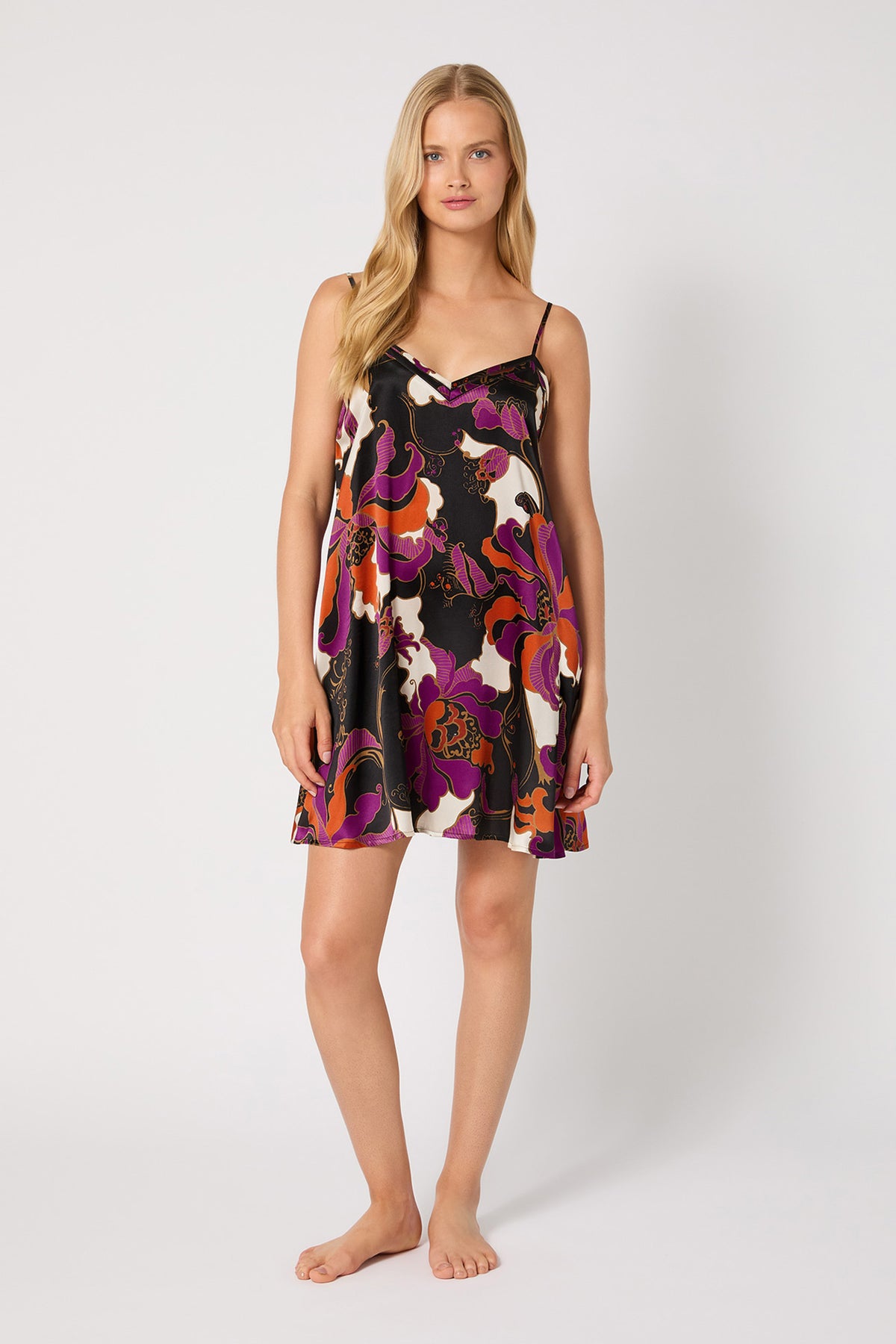 A woman stands barefoot against a plain white background, wearing the Silk Chemise Nightgown - Fall Floral, a sleeveless A-line slip dress with an abstract floral print in black, pink, orange, and purple.