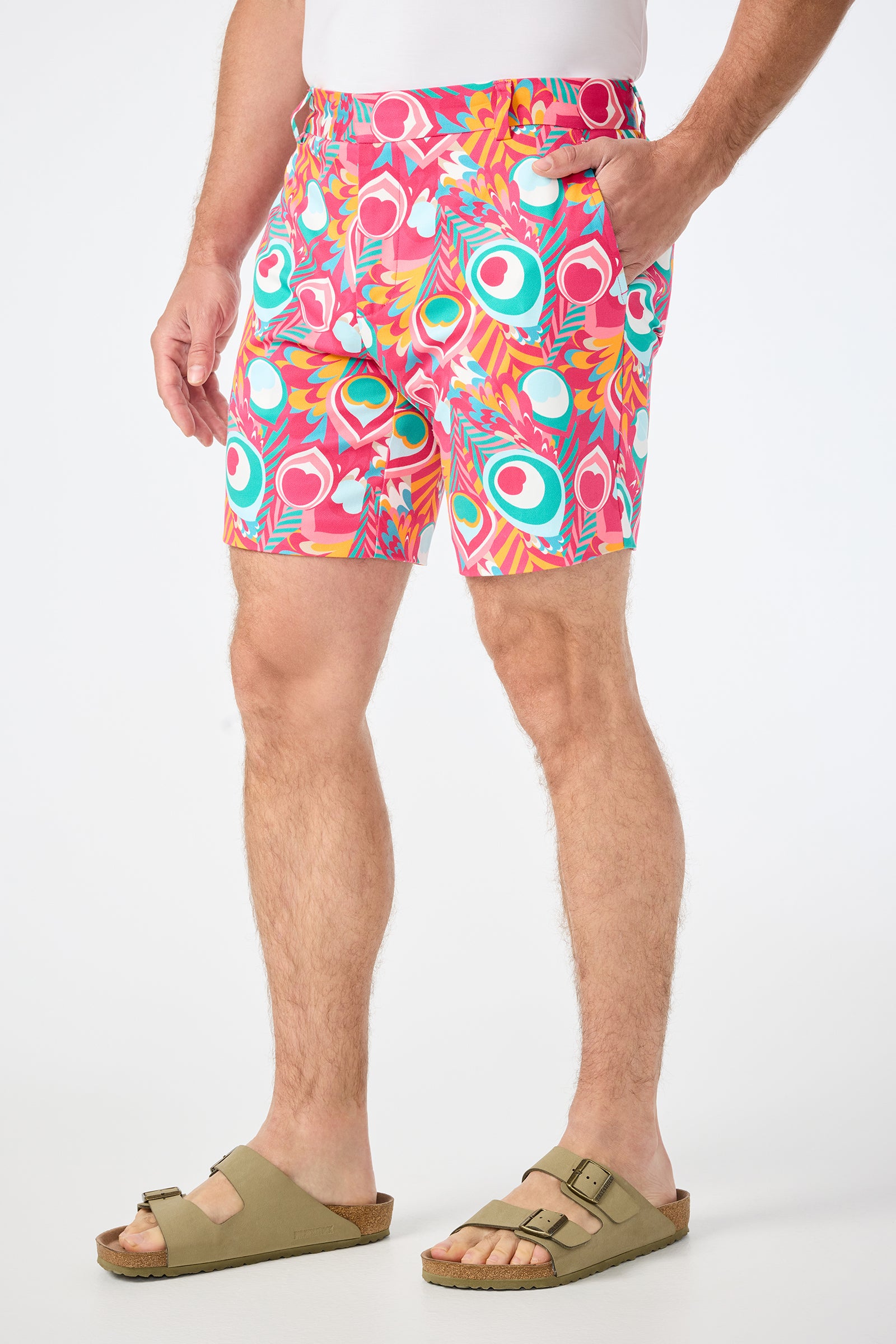A man stands with his hands in his pockets against a plain white background, wearing a white shirt, beige sandals, and the Lawrence Short - Dramont Herringbone featuring pink, teal, and orange patterned shapes.