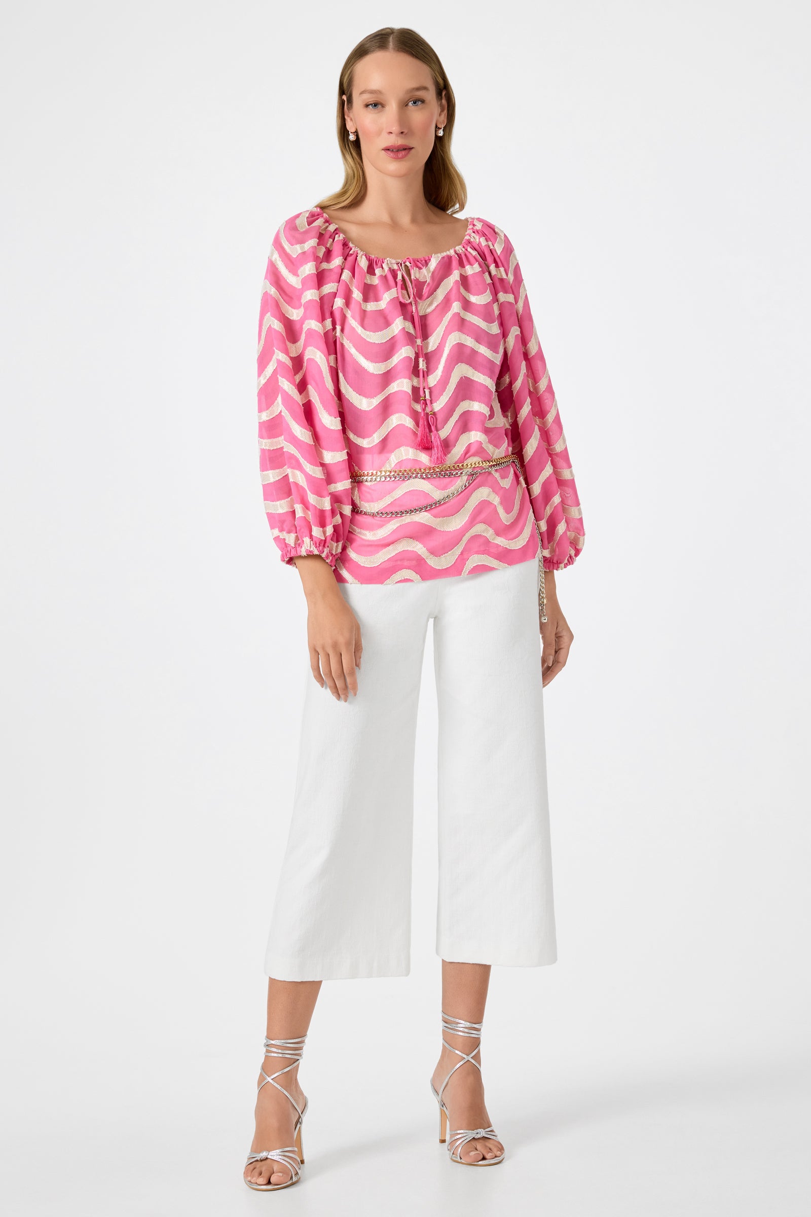 A woman stands against a white background wearing the Sandia 2 Blouson Top - Pinku, featuring a pink and white wavy pattern with three-quarter sleeves, paired with white wide-legged cropped pants and silver strappy heels.
