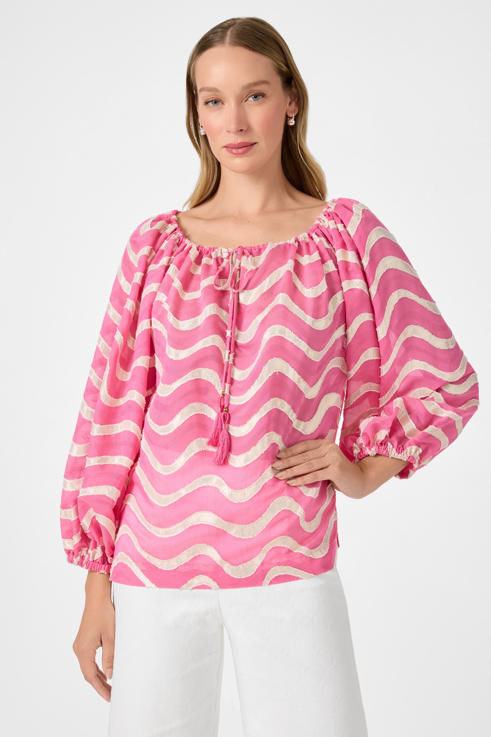 A woman poses against a plain light background wearing the Sandia 2 Blouson Top in Pinku, featuring pink and white wavy stripes, balloon sleeves, and a tasseled neckline tie, paired with white pants.