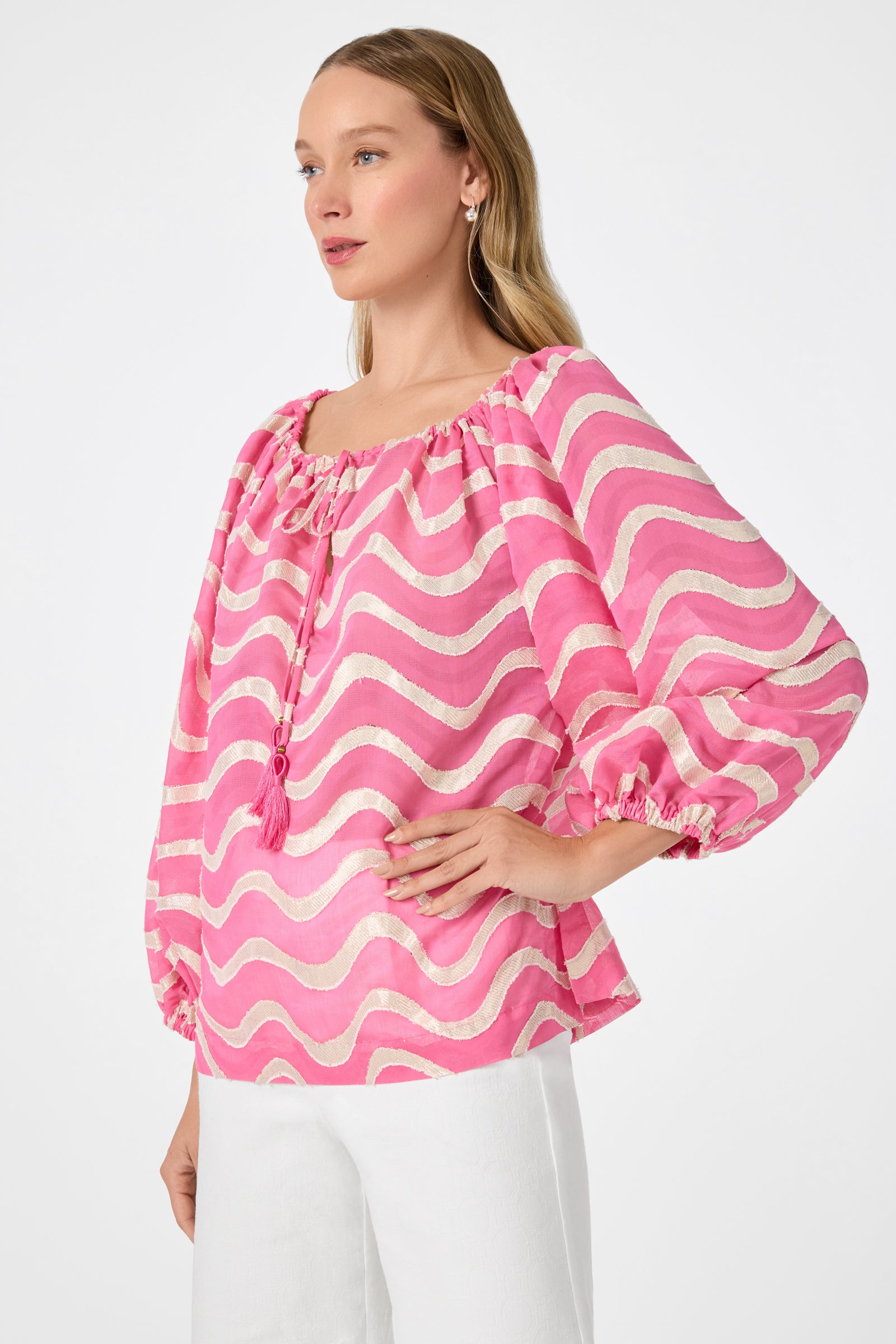 A woman stands with one hand on her hip against a plain background, wearing the Sandia 2 Blouson Top - Pinku, a loose, long-sleeved pink and white wavy-striped blouse with a tie neckline, paired with white pants.