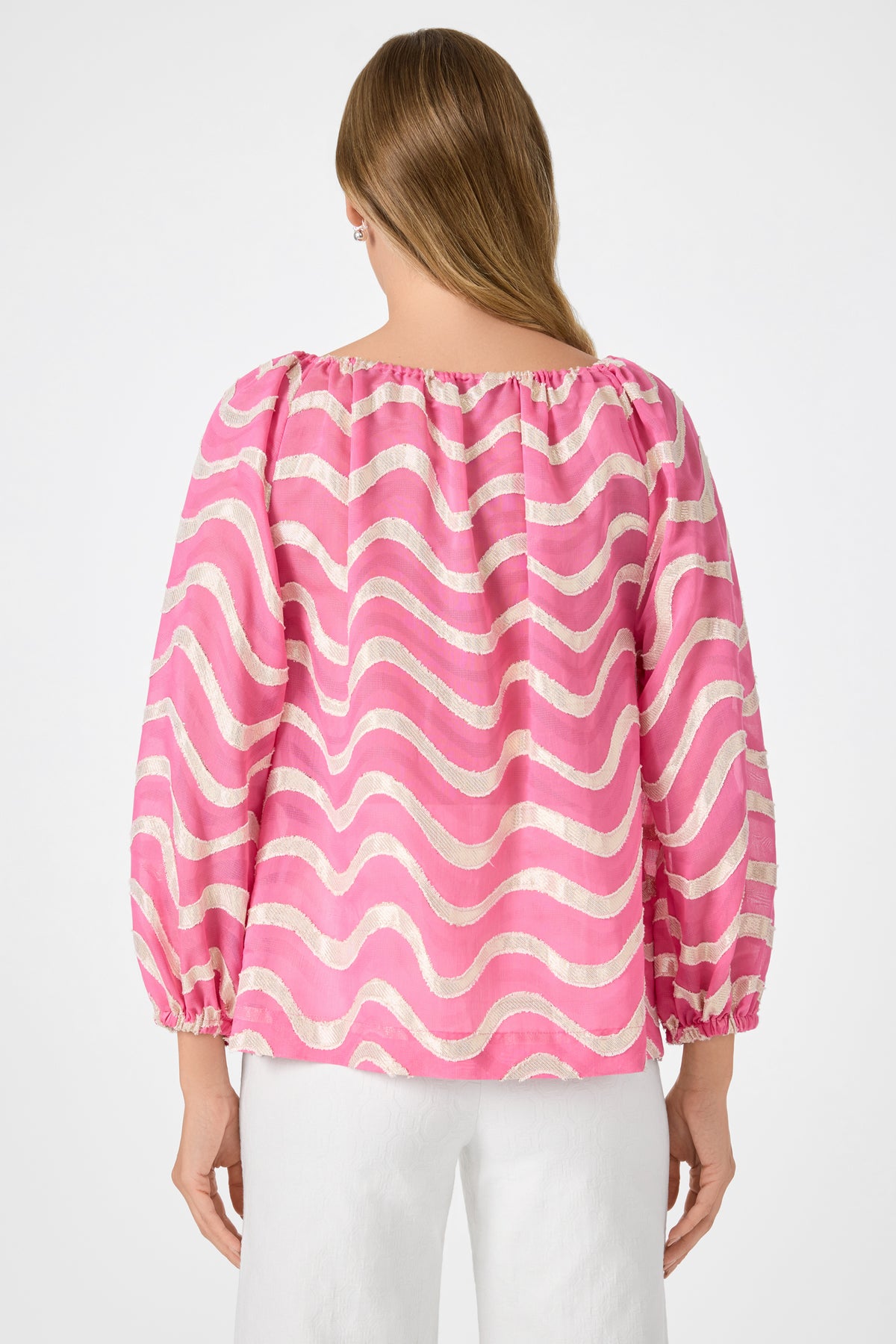 A person with long, light brown hair is seen from the back wearing the Sandia 2 Blouson Top - Pinku, a loose-fitting pink and white wavy-striped blouse with balloon sleeves, paired with white pants against a plain light background.