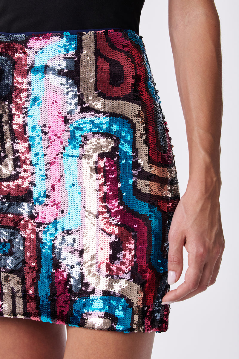 A close-up of a person wearing the Rico Skirt, a sequined mini with an abstract pattern in pink, blue, black, and gold. Their right hand rests by their side against a plain background.