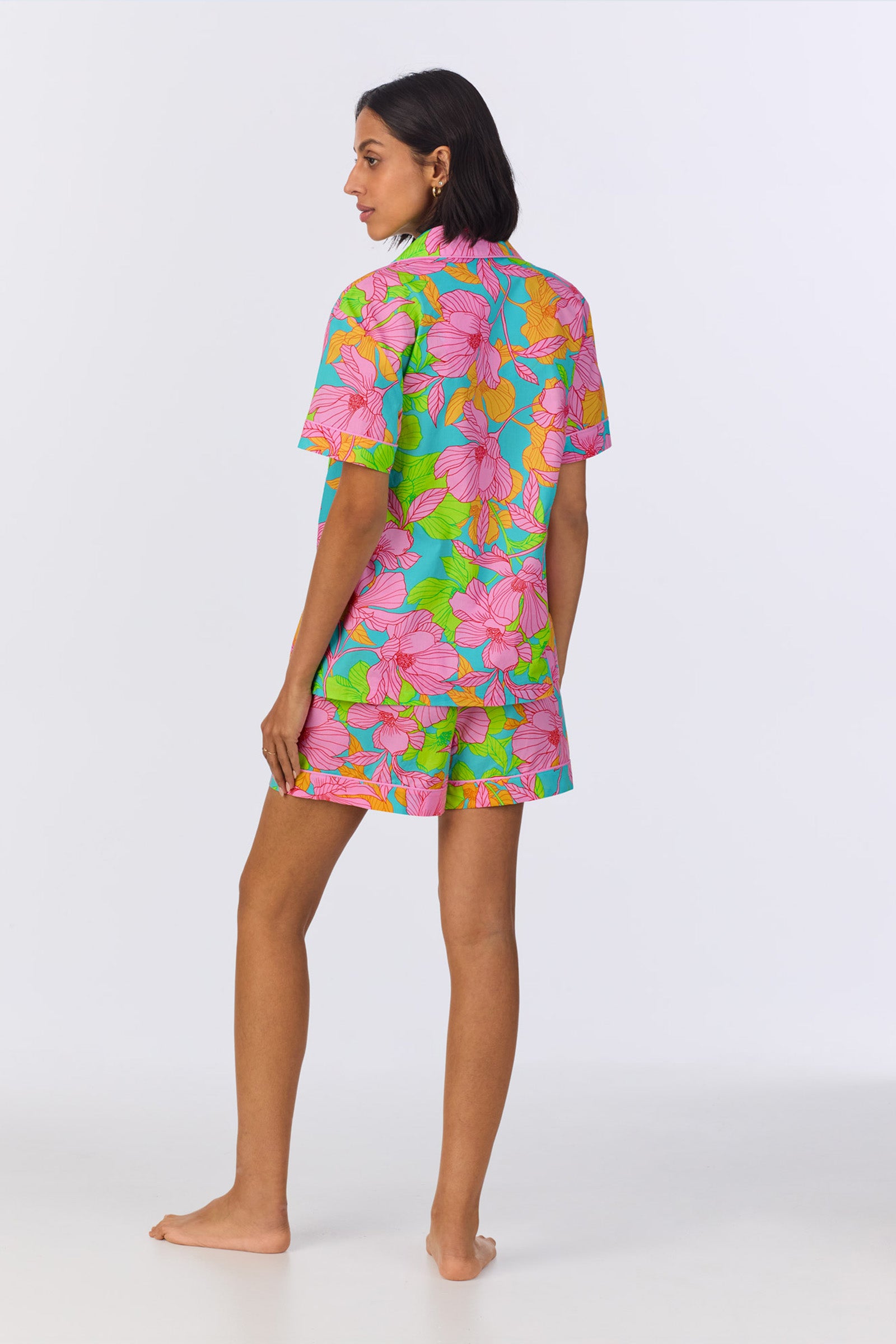 A person stands barefoot, facing away, wearing the Shorty PJ Set - Linear Floral Poplin—a matching floral-patterned short-sleeve shirt and shorts in pink, green, orange, and blue tones—on a plain white background.