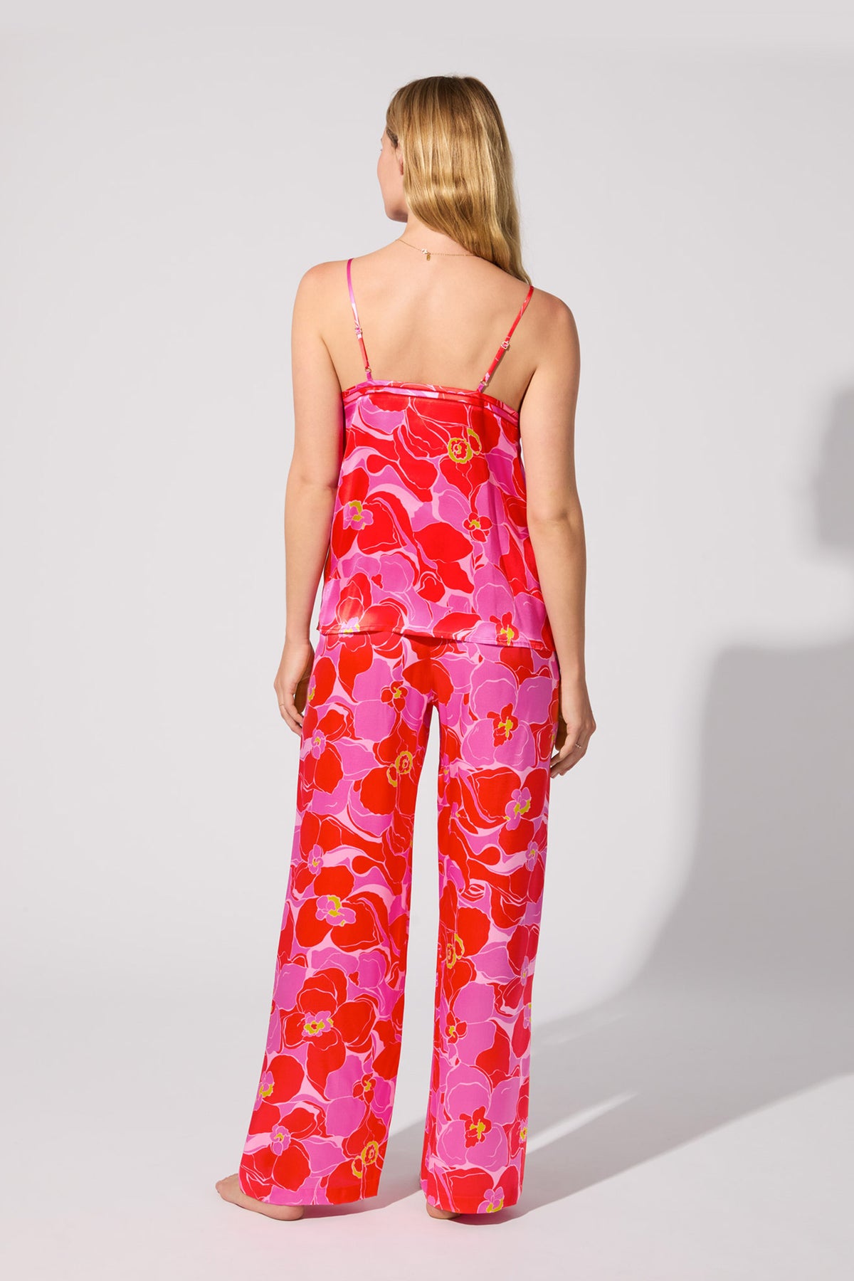 A woman with long blonde hair faces away, wearing the Silk Cami PJ Set - Winter Orchids, featuring a strappy top and loose pants with a bold pink, red, and purple floral print. The background is plain light gray.