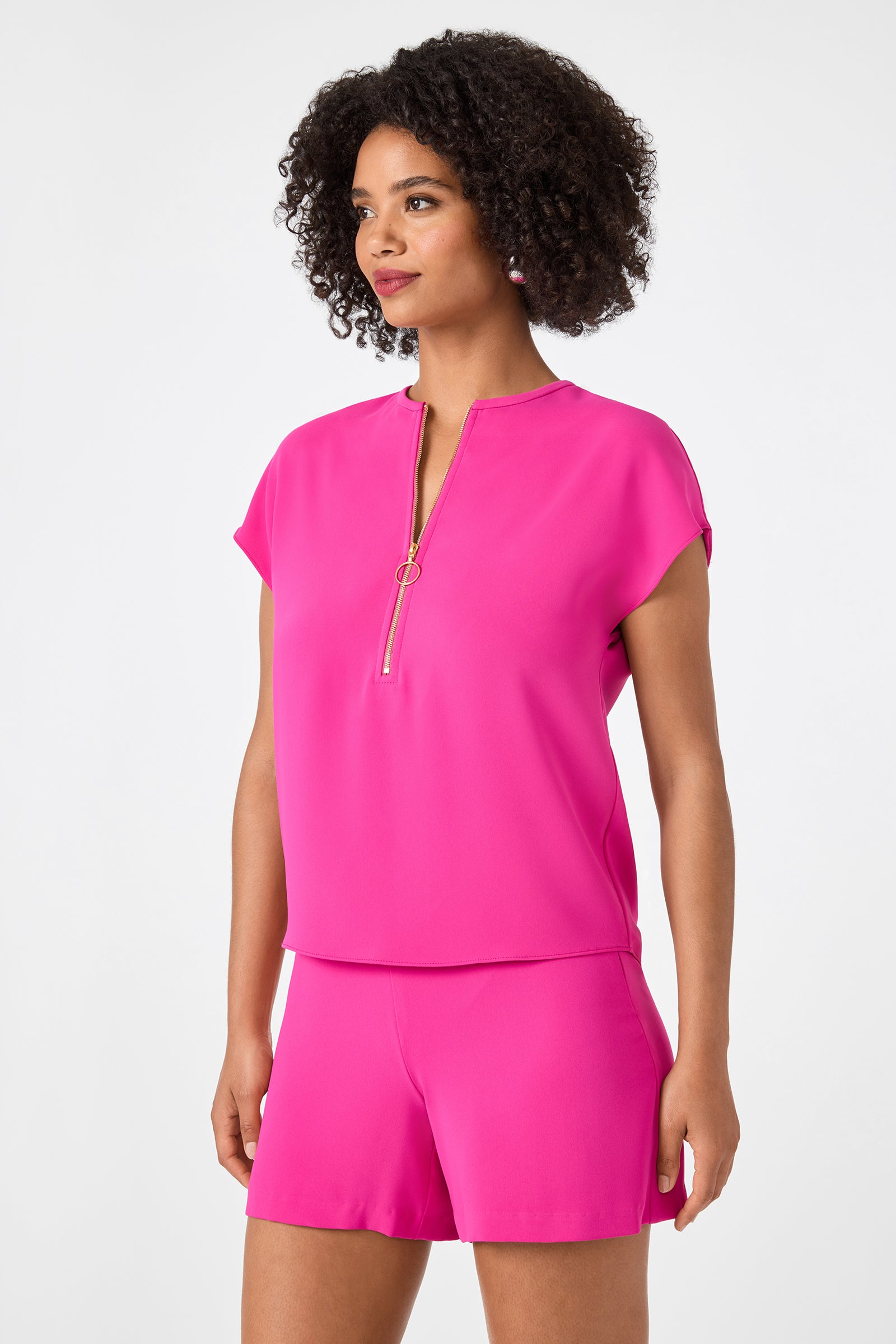 A woman with curly hair wears the Hasil Top - Double Georgette in bright pink, featuring short sleeves and a front zipper, paired with matching pink shorts against a plain light background.