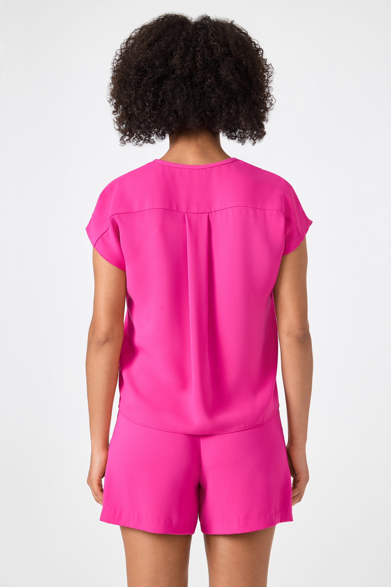 A person with curly hair is facing away from the camera, wearing the Hasil Top - Double Georgette in bright pink and matching shorts against a plain white background.