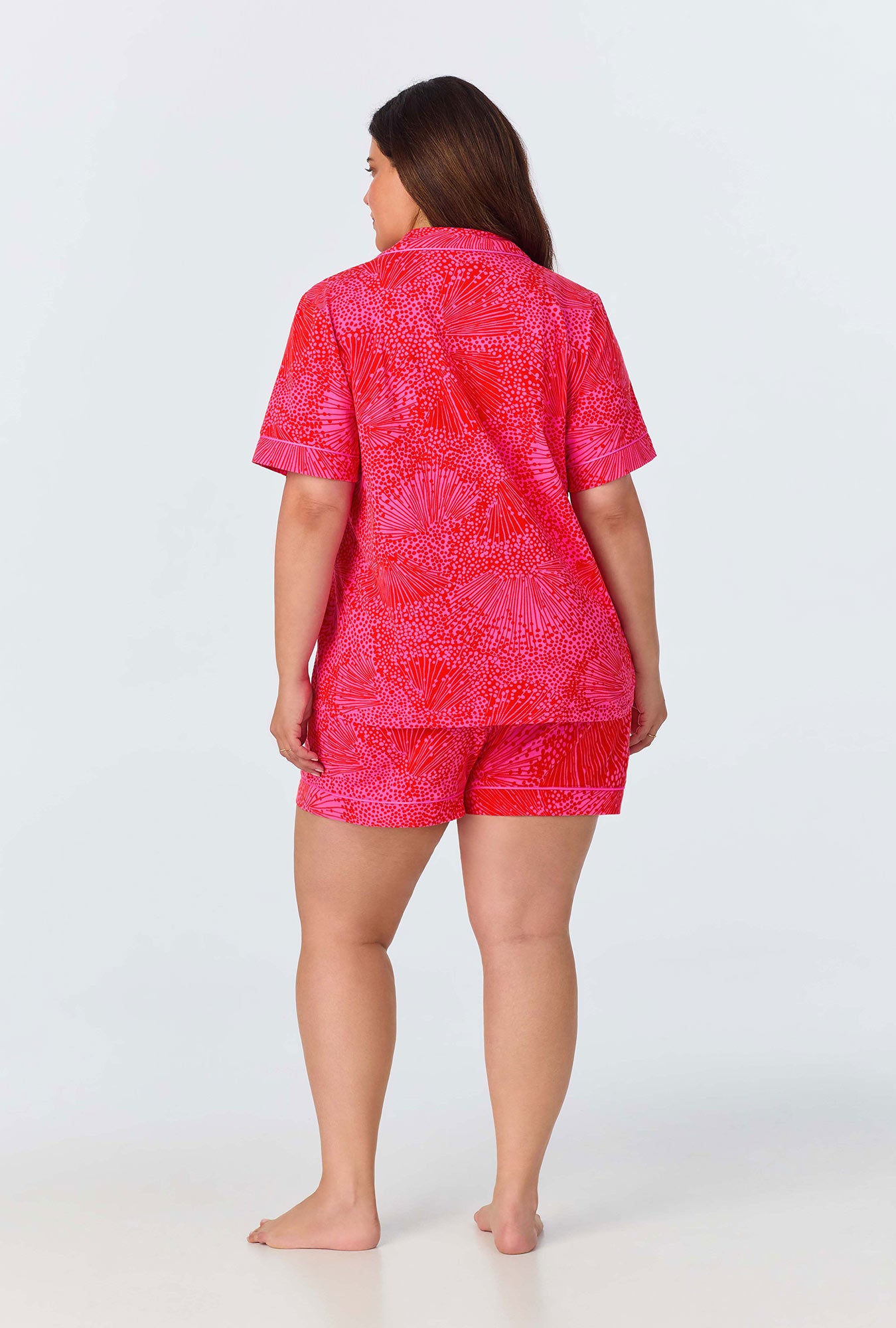 A woman stands barefoot, facing away from the camera, wearing the Starburst Womens Short-Sleeve Shorty Jersey PJ Set—featuring a red and pink pattern—against a plain light background.