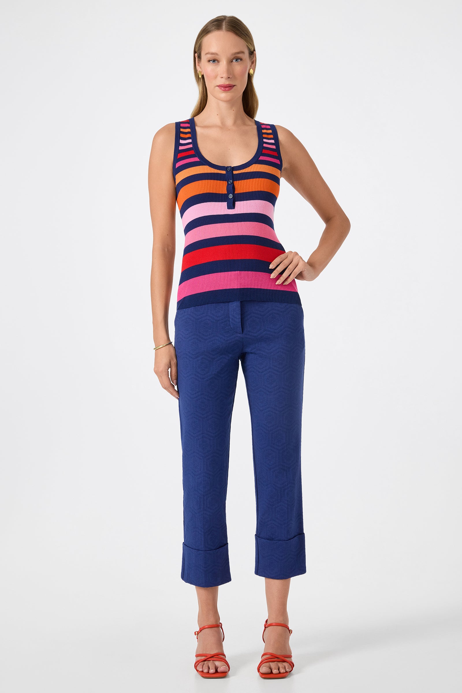 A woman with straight, light brown hair and a neutral expression wears the Arles Scoop Neck Tank—a sleeveless, multicolored striped top—paired with blue cropped pants and red strappy sandals against a plain background.
