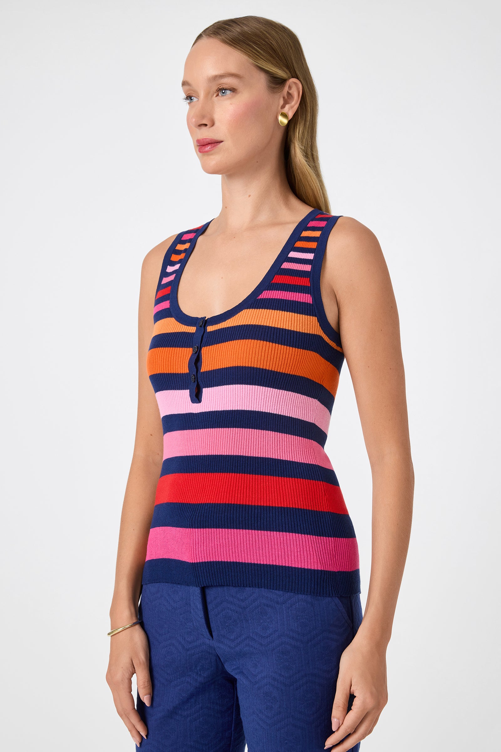 A woman with long light brown hair poses against a plain light background wearing the Arles Scoop Neck Tank— a sleeveless, knit top with horizontal stripes in navy, orange, red, pink, and purple—paired with blue pants.