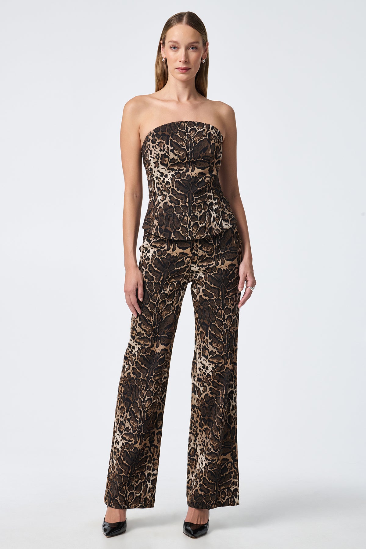 A woman stands facing forward in the Juji Bustier, a strapless, fitted top, paired with high-waisted, flared leopard print pants. She has straight shoulder-length hair and black heels against a plain white background.