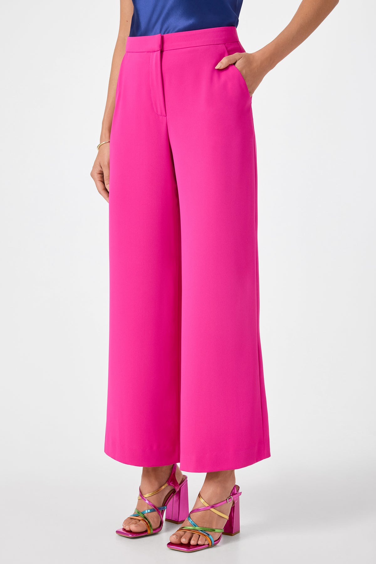 Wearing Verity 3 Wide-Leg Pants in bright pink, paired with a blue top and colorful strappy heels, a person stands with one hand in their pocket against a white background.