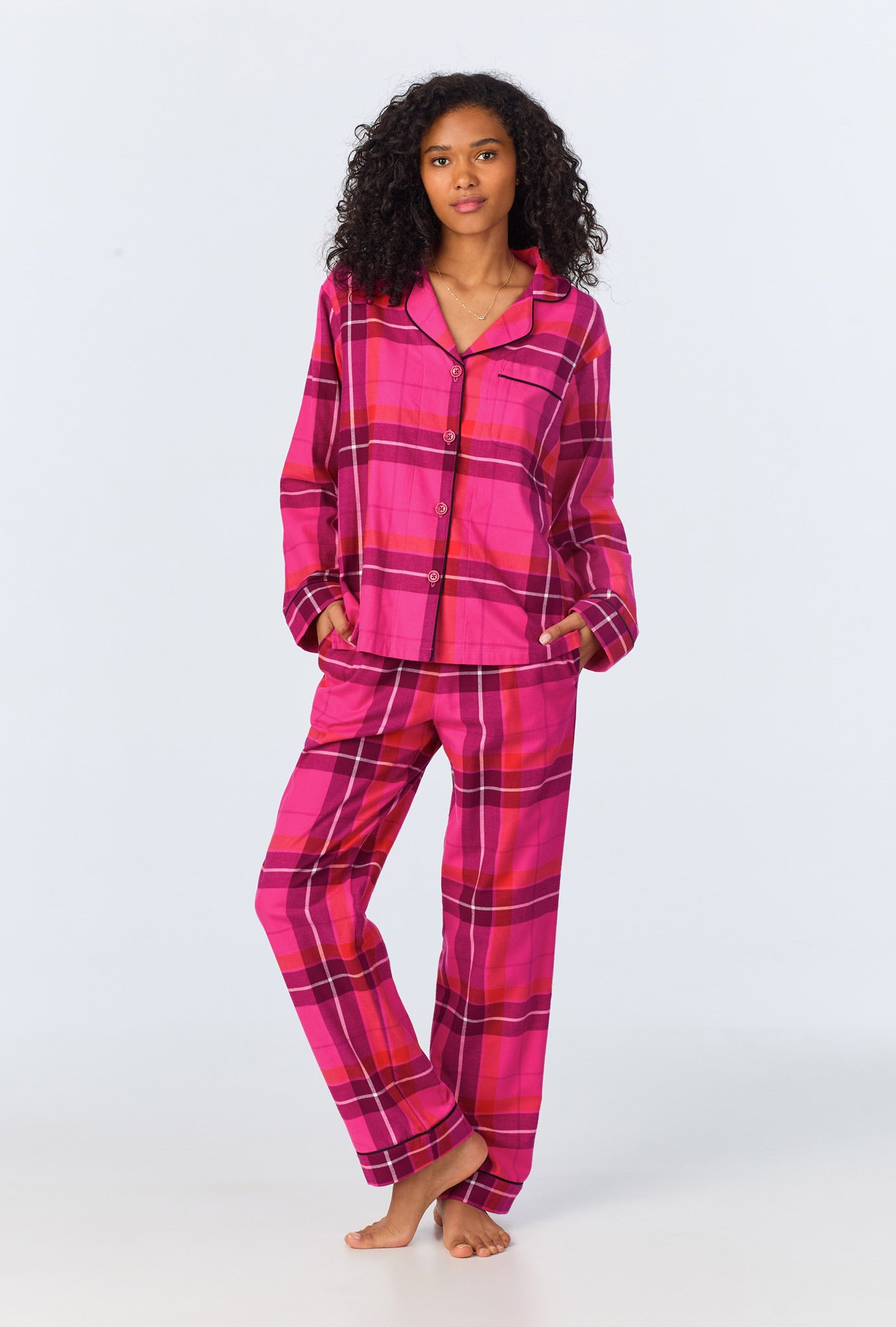 A woman stands barefoot wearing the Aurora Plaid Womens Long Sleeve Long Pant Flannel PJ Set, featuring a collared button-up top and plaid pants with cozy pockets in bright pink and magenta.