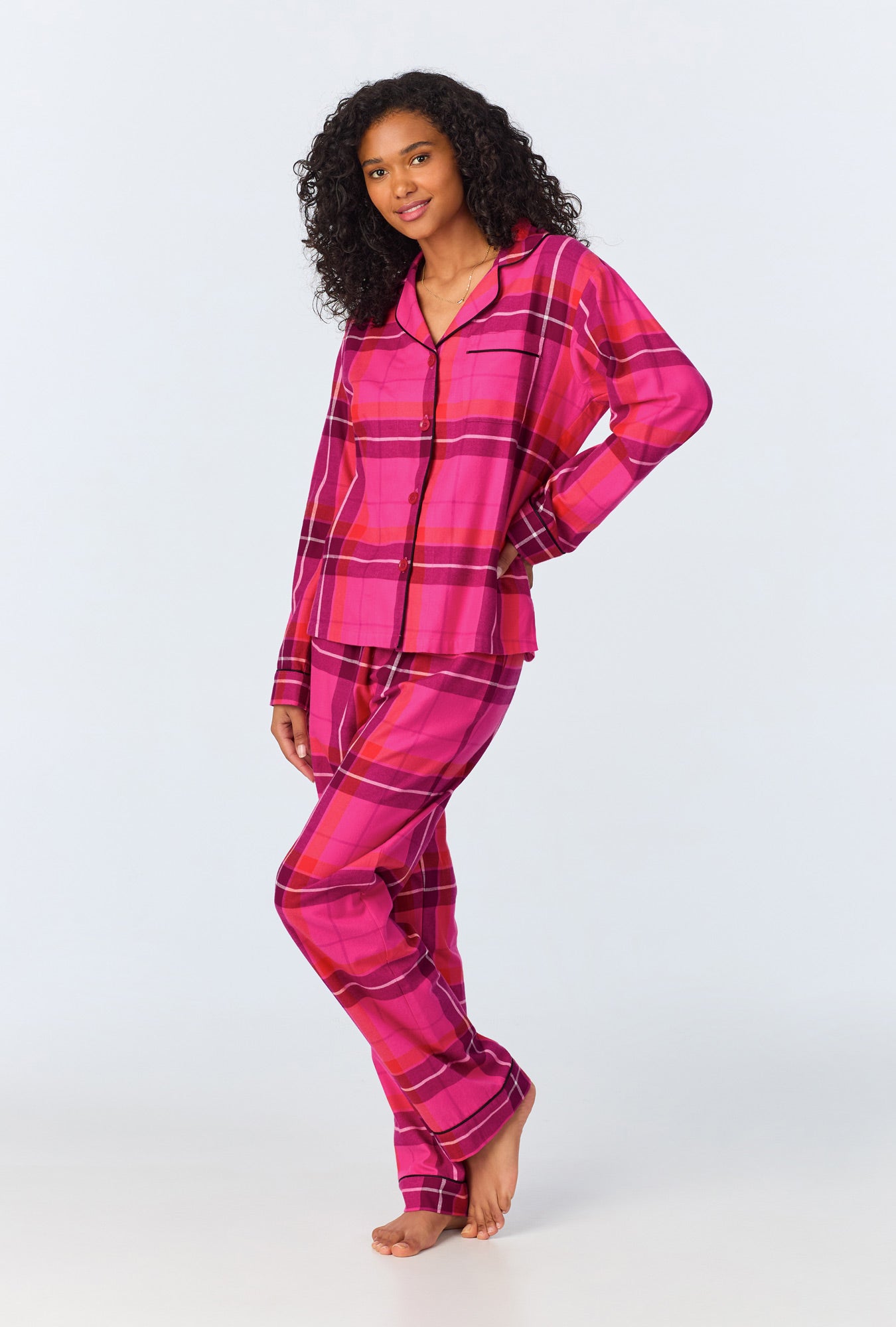 A woman with curly hair stands barefoot, smiling slightly in the Aurora Plaid Womens Long Sleeve Long Pant Flannel PJ Set—hot pink and magenta plaid, button-up top and pants. One arm is bent at her side against a plain background.