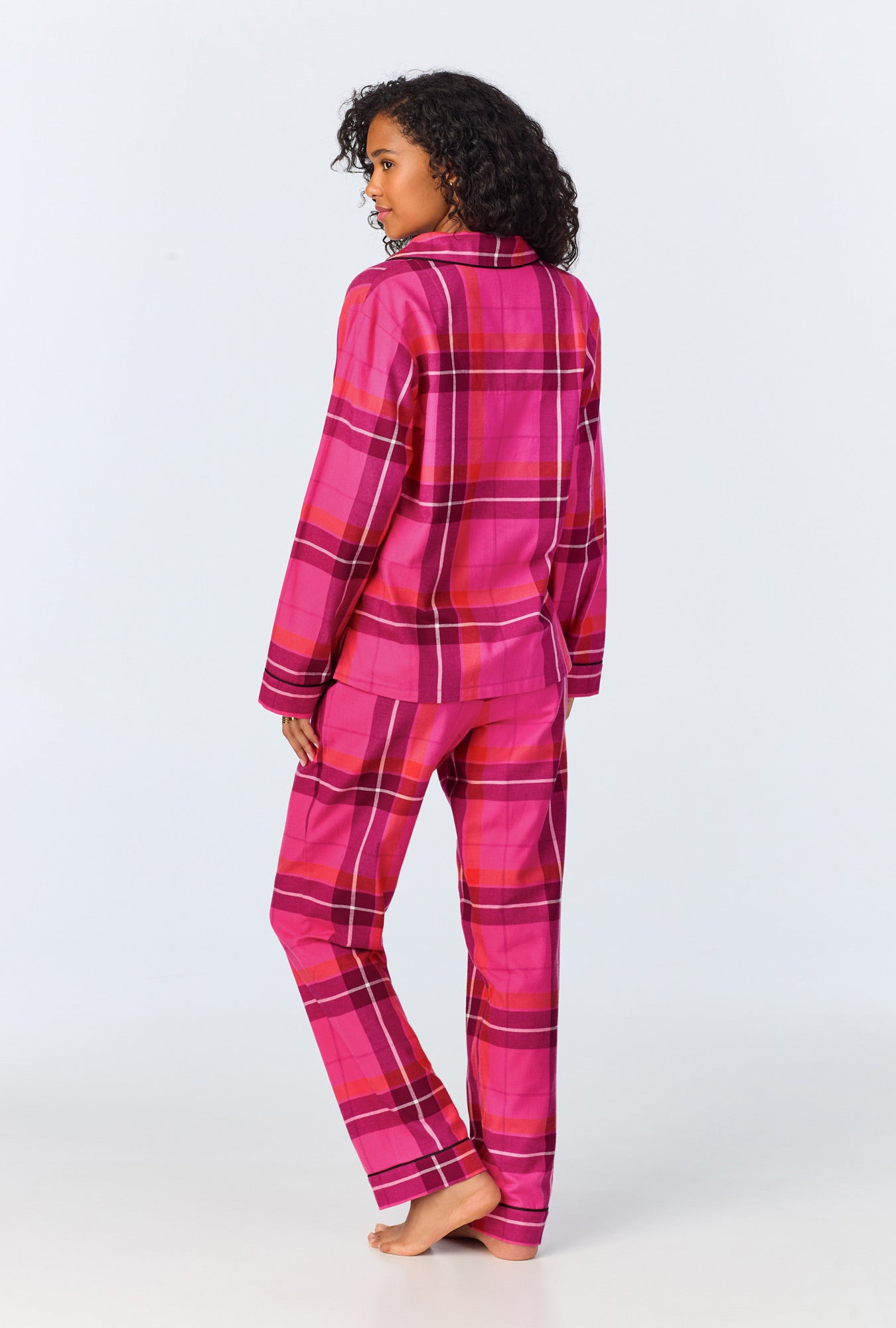 A woman stands barefoot, facing away, wearing the Aurora Plaid Womens Long Sleeve Long Pant Flannel PJ Set. The pajama set features a vibrant plaid pattern. The background is plain and light-colored.