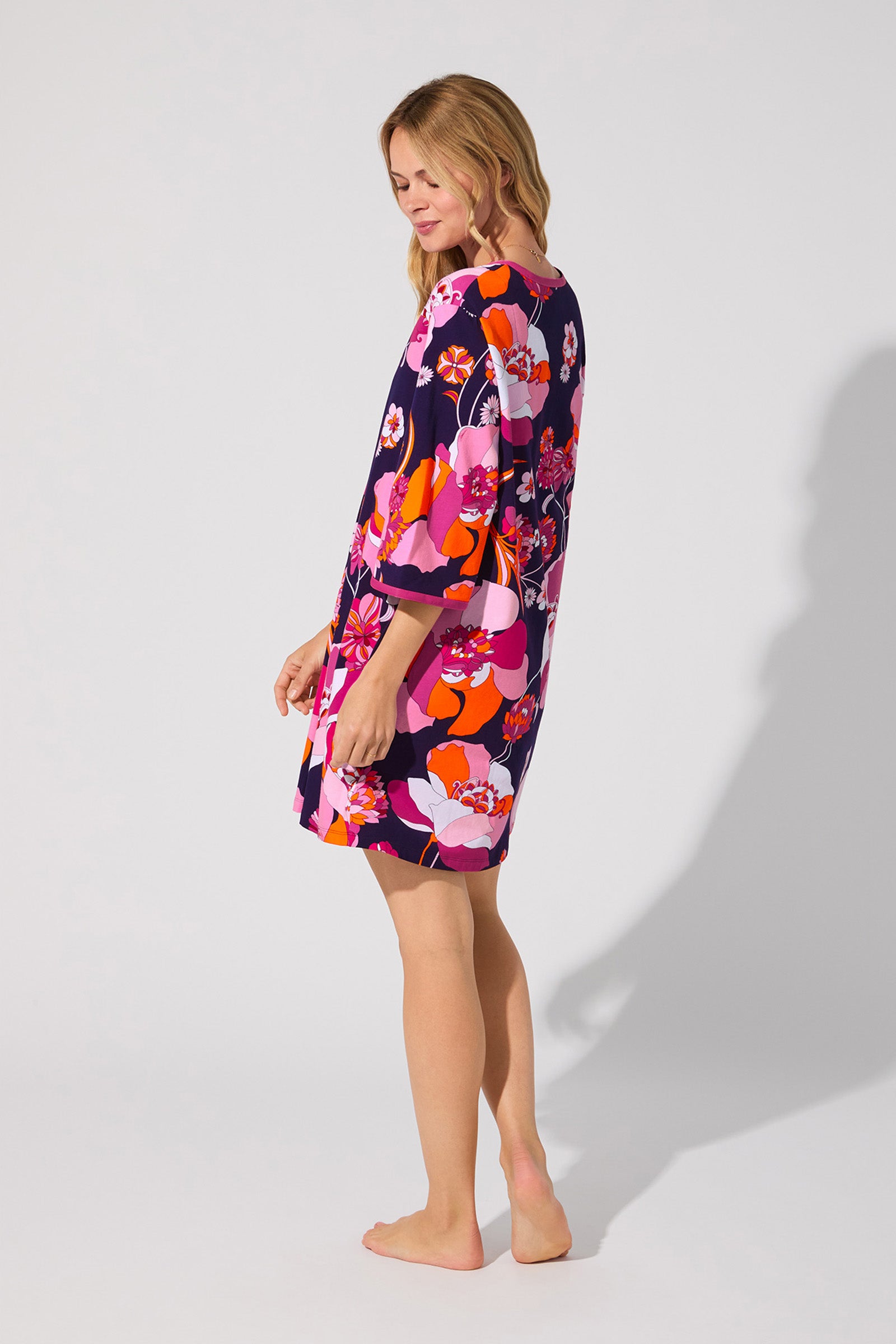 A blonde woman stands barefoot, smiling gently in the Floral Medley Caftan—a loose, knee-length dress with bright pink, orange, red, and purple floral patterns—set against a plain background.