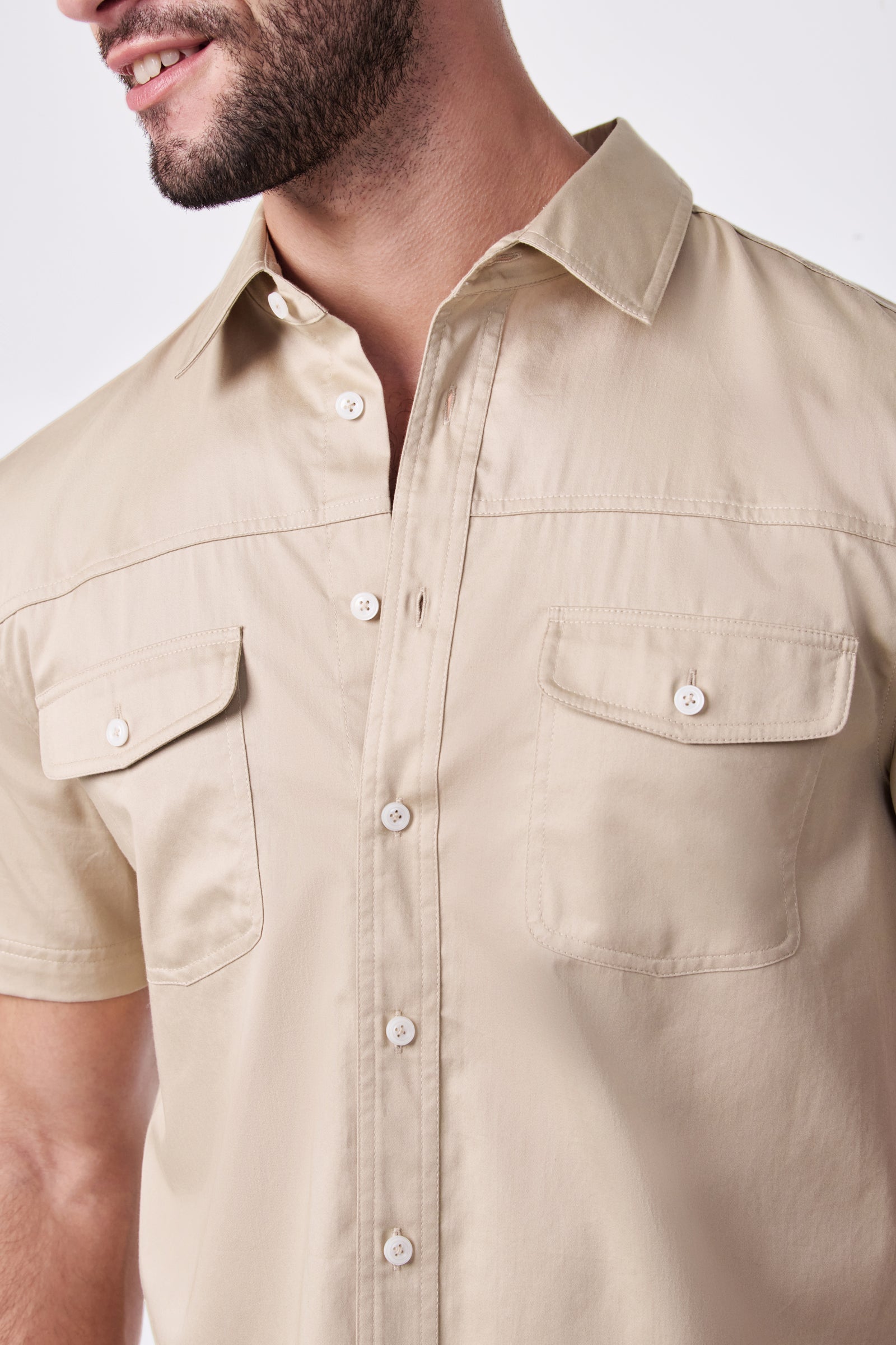 A man wears the Shelton Ss Shirt, a beige short-sleeve button-up with two chest pockets. Cropped at his chin, the photo shows only the lower half of his face and upper torso against a plain background.
