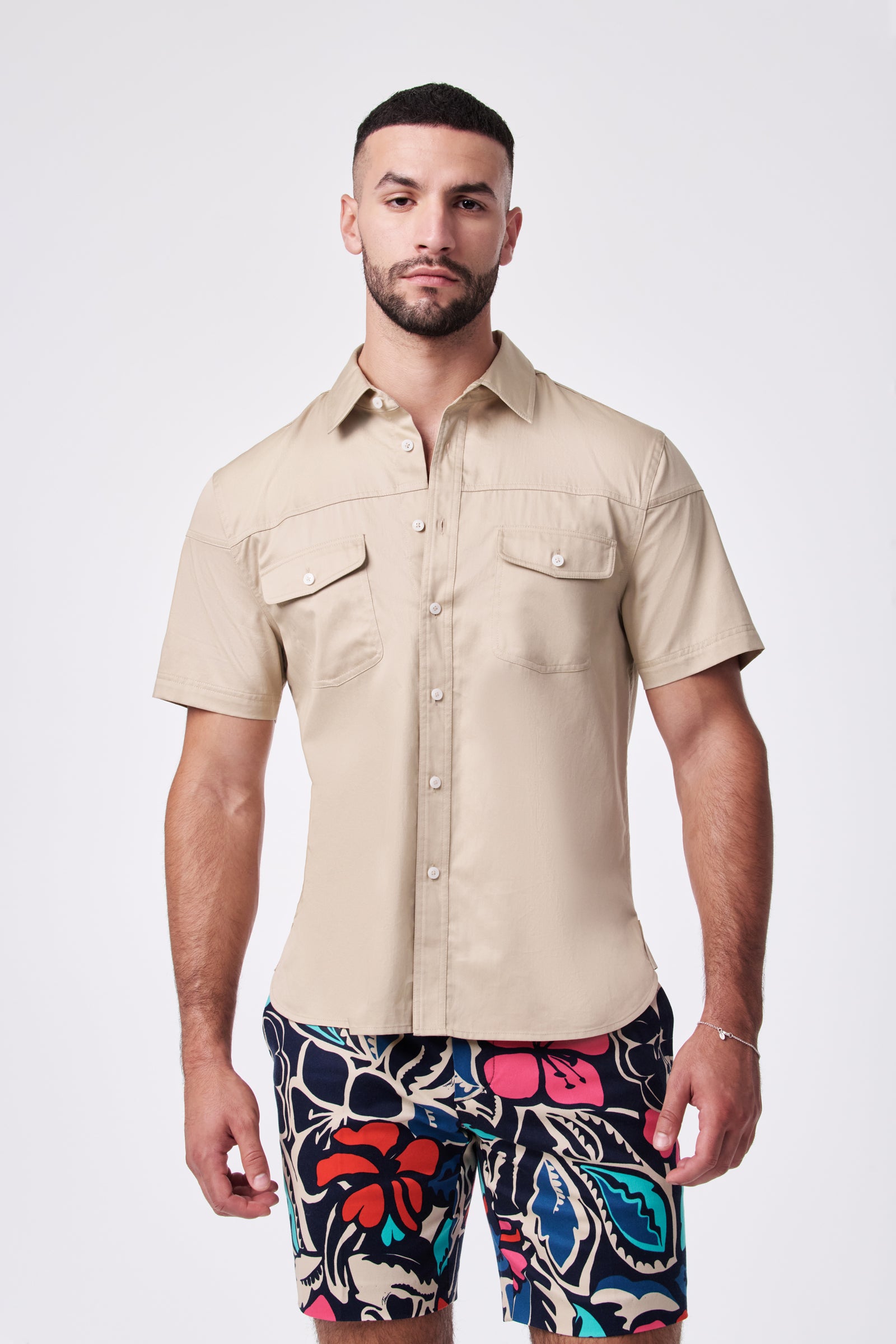 A man faces forward against a plain white background, wearing the beige Shelton Ss Shirt with front pockets and colorful patterned shorts.