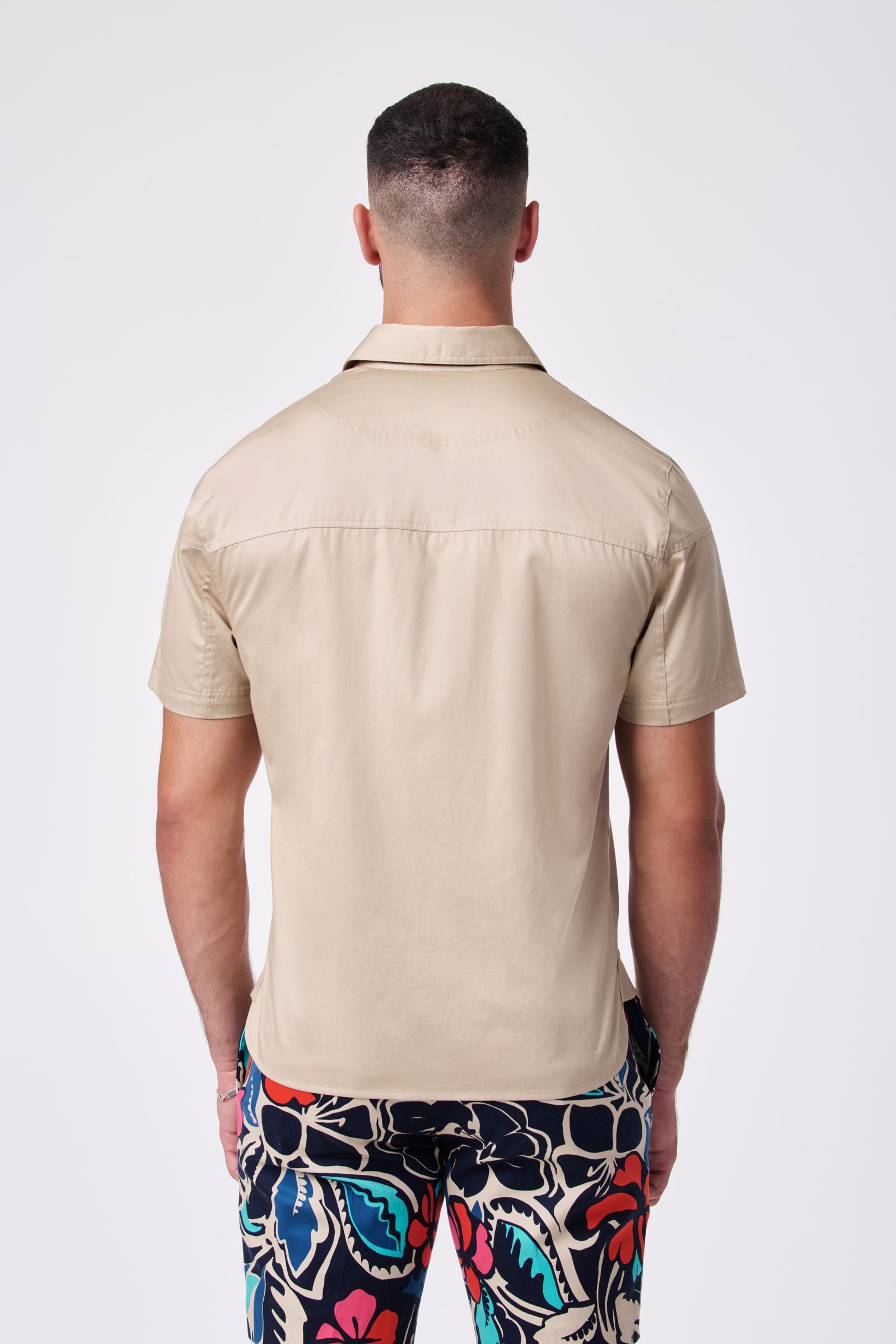 A man facing away from the camera wears the Shelton Ss Shirt, a beige short-sleeved top, with colorful floral-patterned pants. The background is plain white.